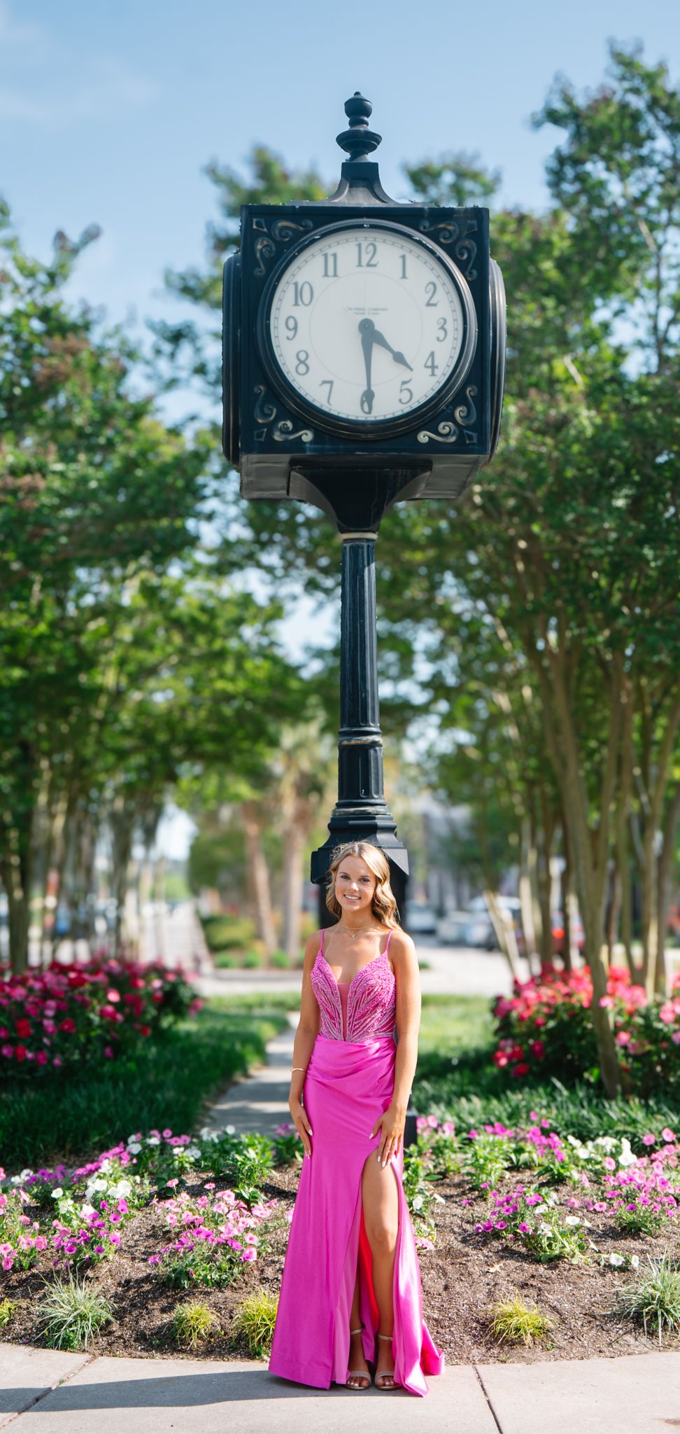 Young woman in hot pink formal gown standing beneath a decorative street clock in a garden.