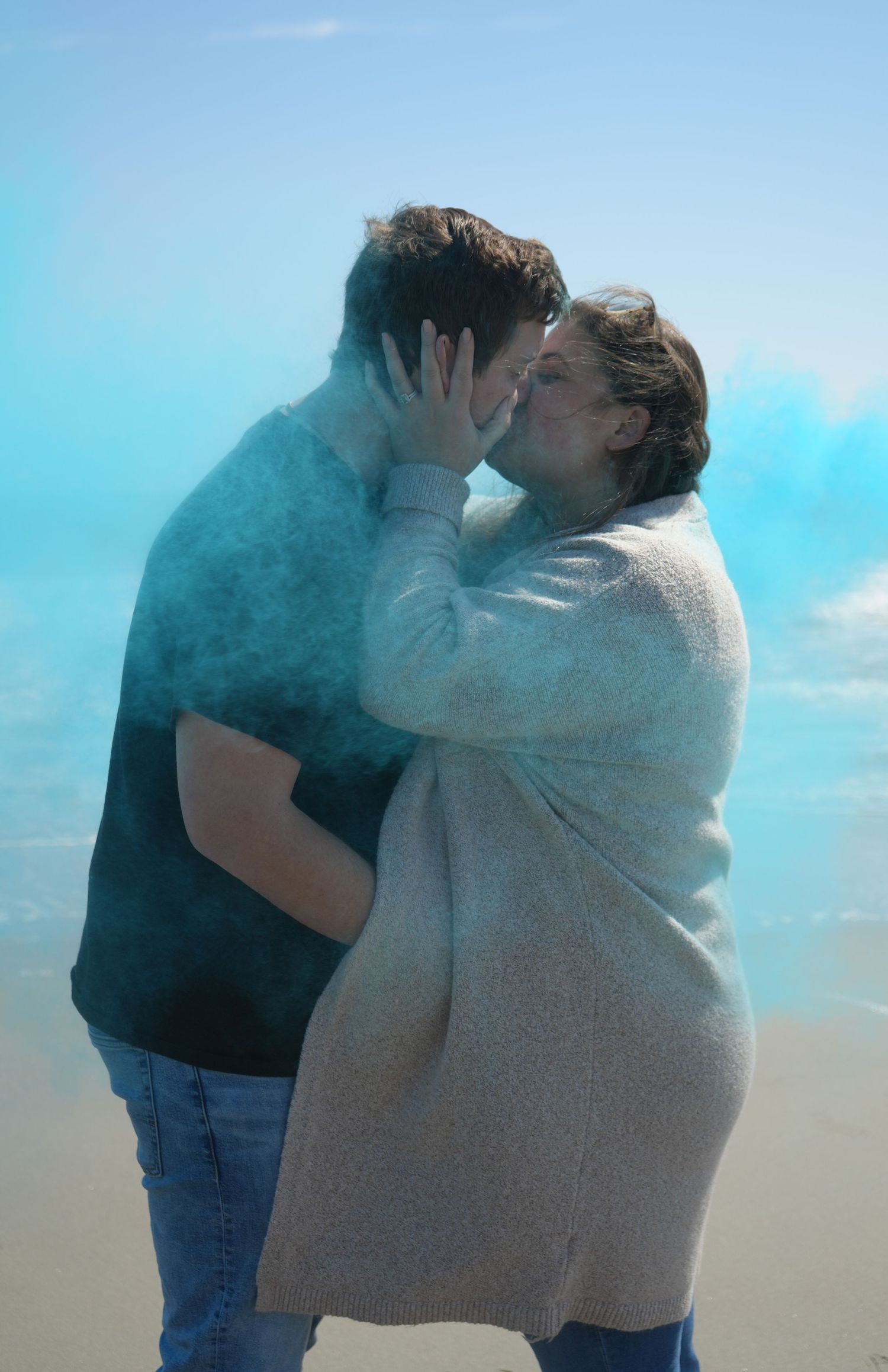 Couple sharing a kiss surrounded by blue gender reveal smoke powder outdoors under a bright sky.