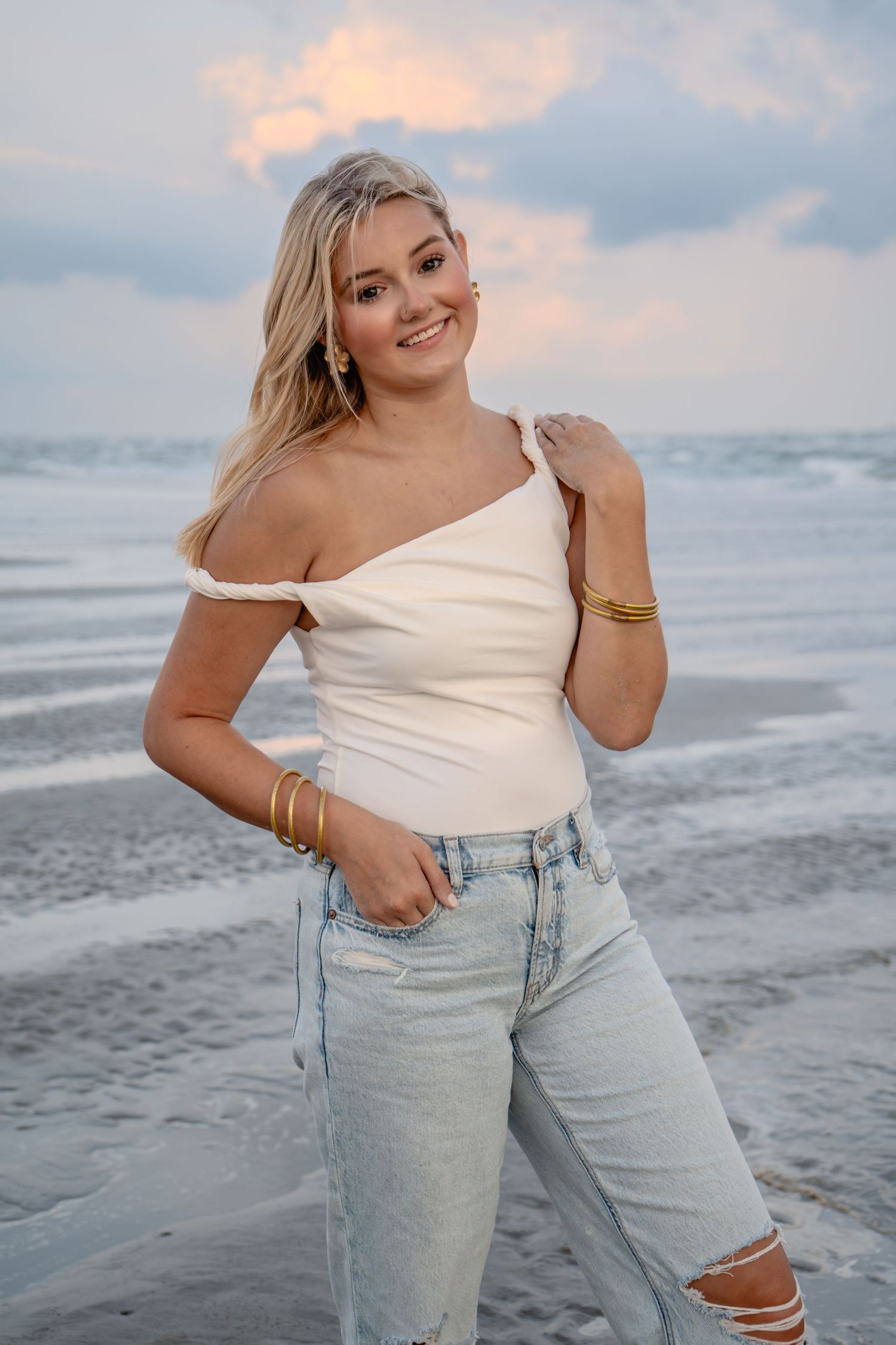 Smiling blonde woman in white off-shoulder top and ripped jeans posing on a beach at sunset.