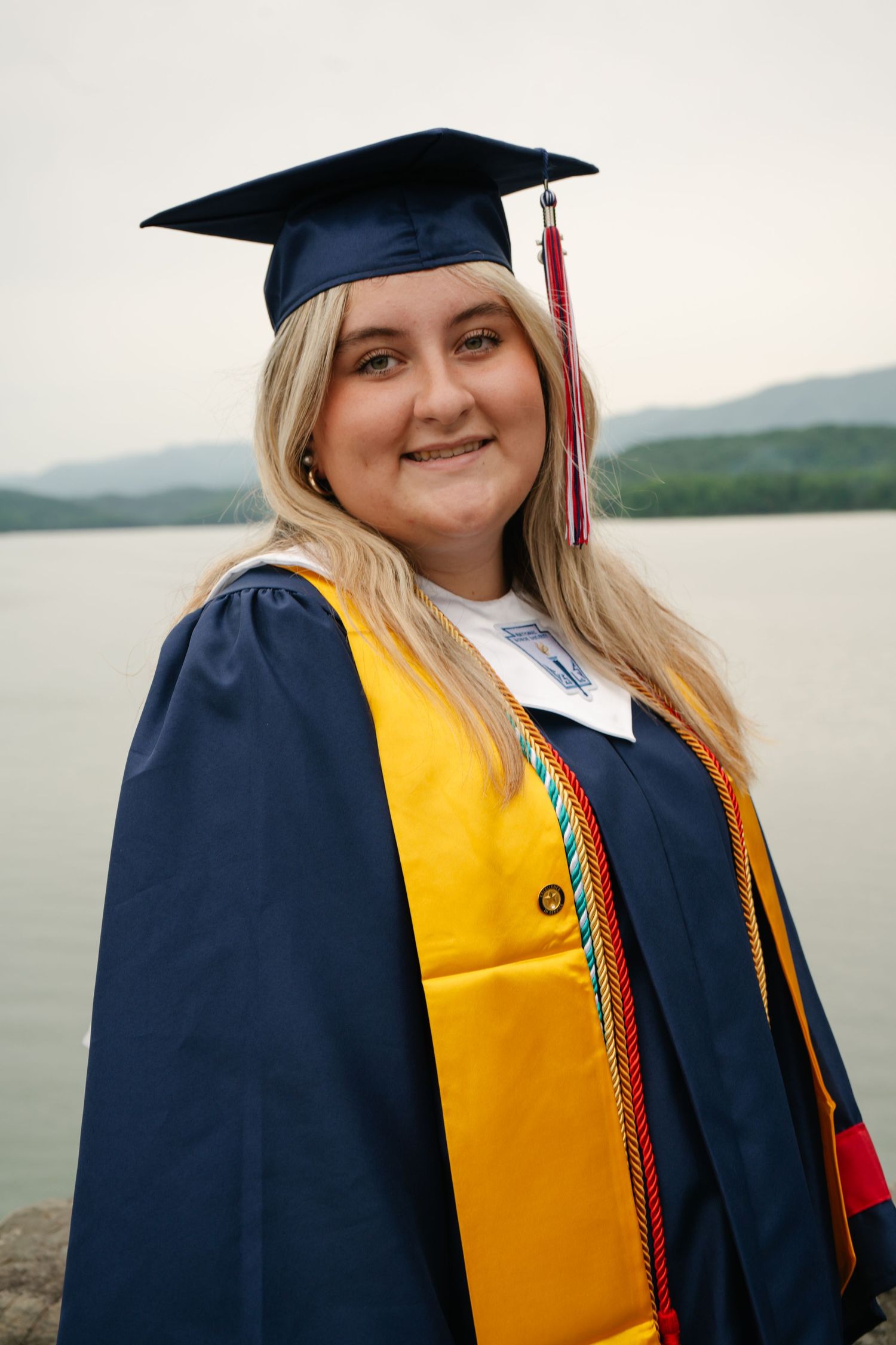 Smiling female graduate in navy and gold cap and gown poses outdoors near a lake with mountain views.