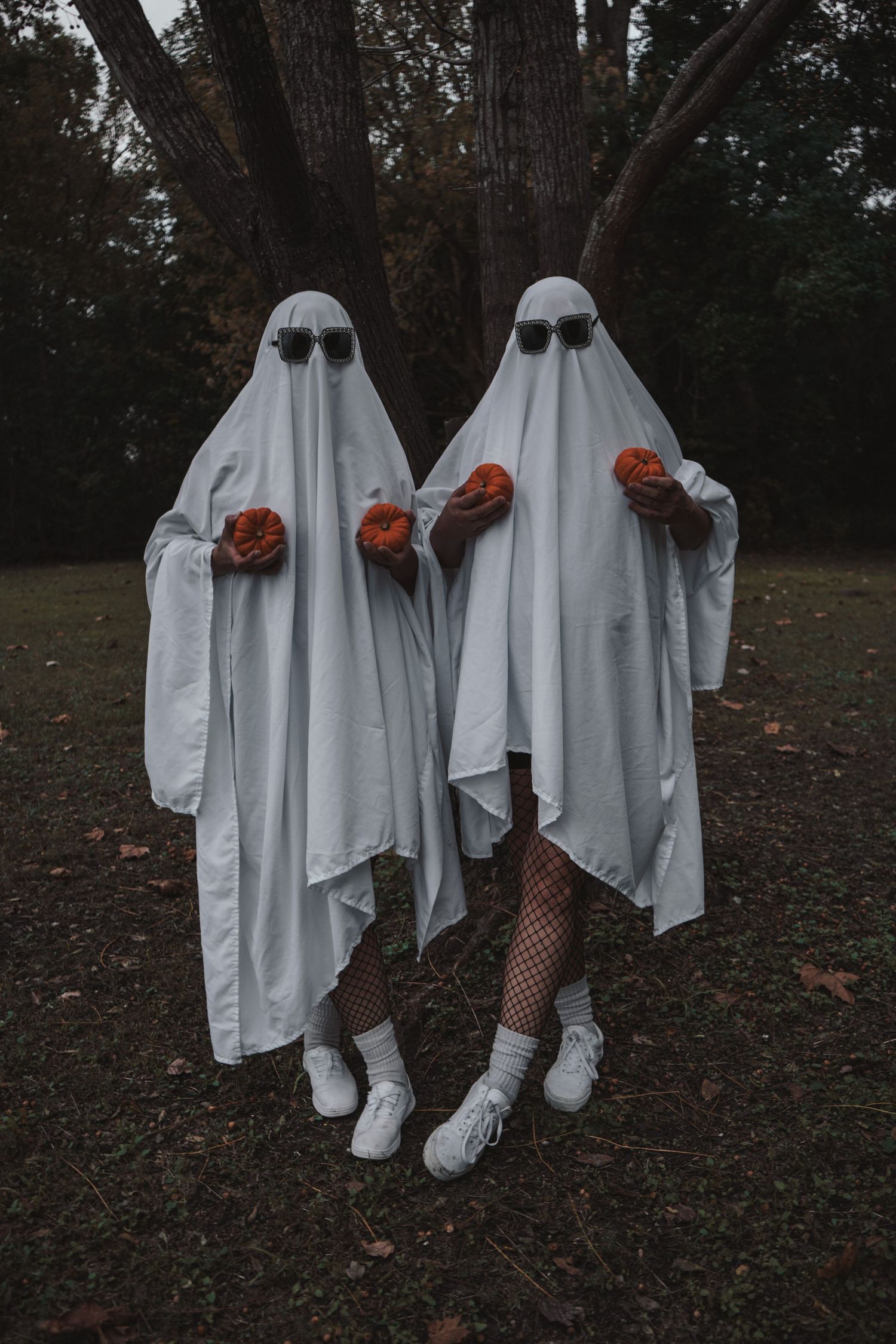 Two people in white ghost Halloween costumes holding pumpkins outdoors.