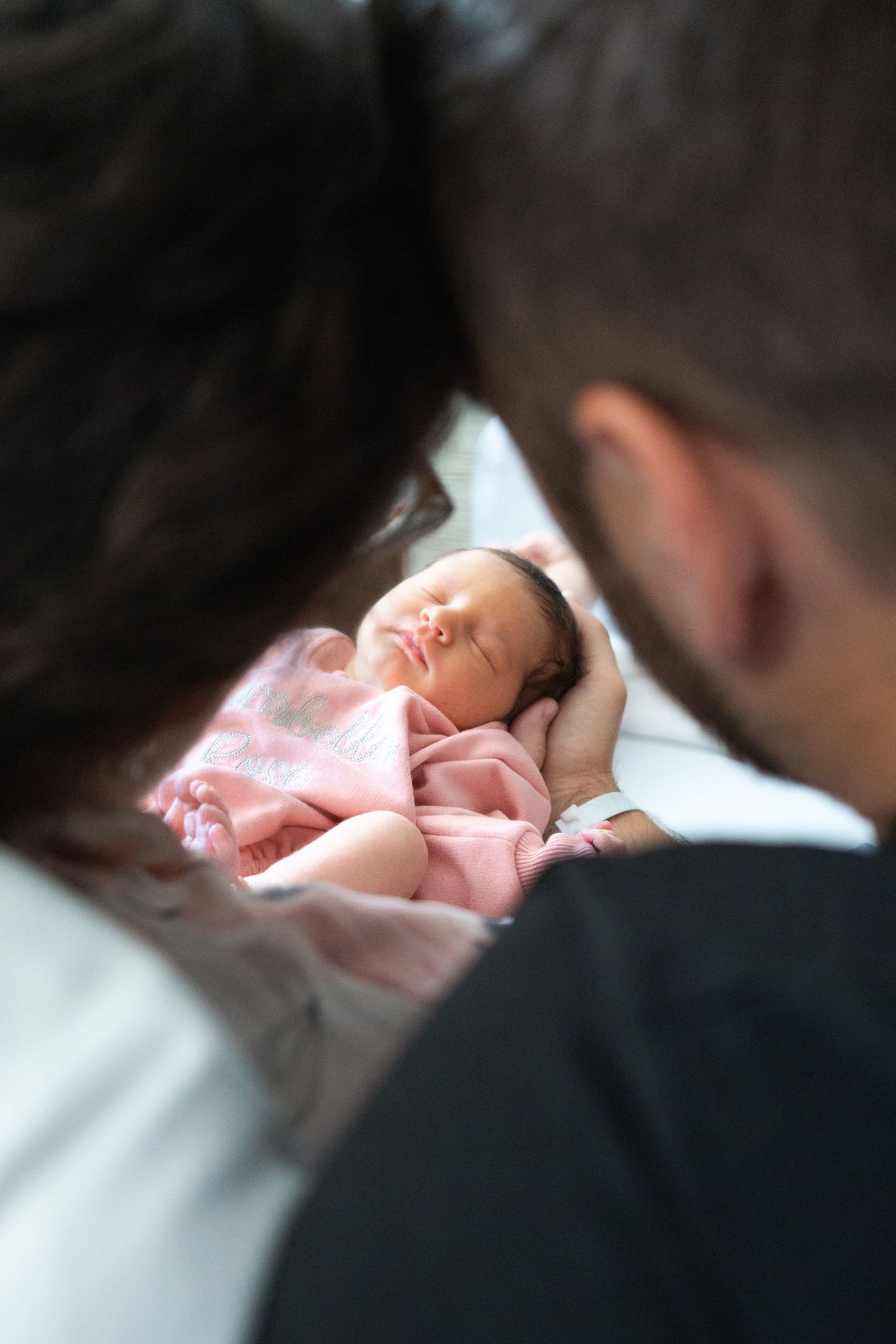 New parents gazing lovingly at their sleeping newborn baby wrapped in pink blanket.