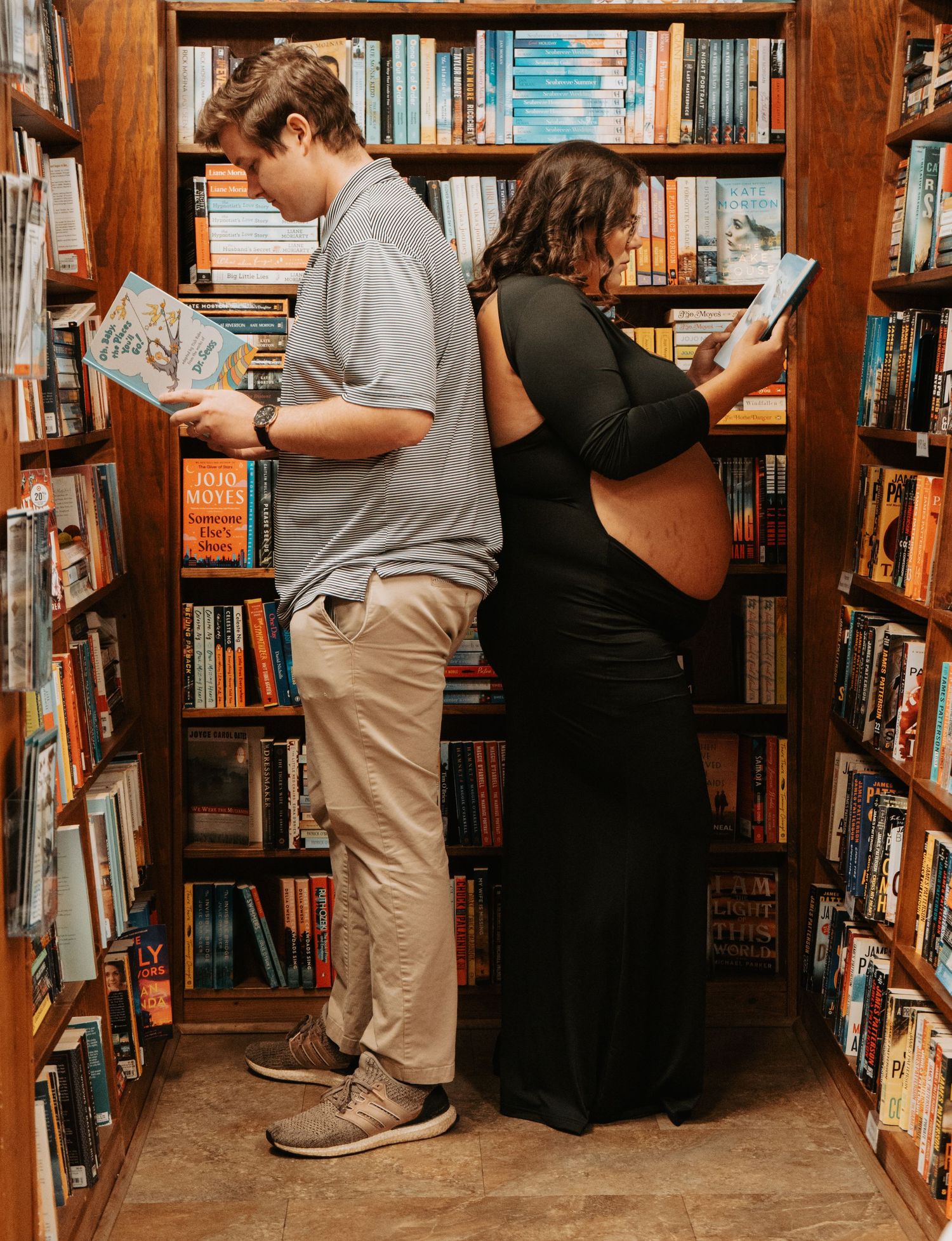 Pregnant woman and teen boy reading books back-to-back in a bookstore aisle.