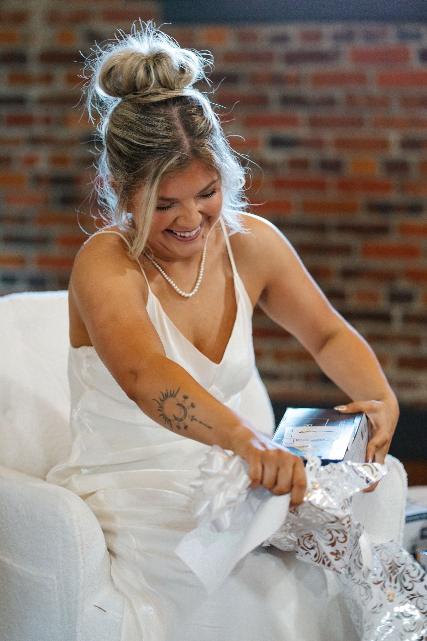Laughing bride in white slip dress opening gifts at bridal shower with exposed brick wall background.