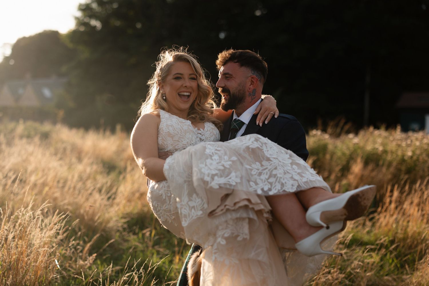 Groom carrying bride in lace wedding dress through golden sunlit field, both laughing joyfully.