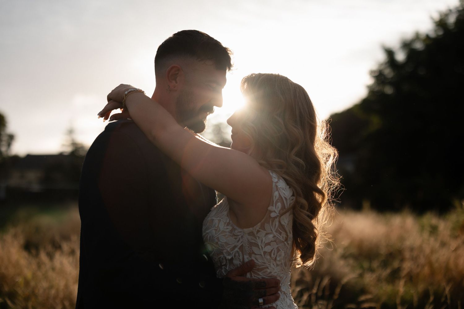 Couple embracing at golden hour sunset in a field, bride in lace gown and groom in dark suit, backlit romantic wedding photo.