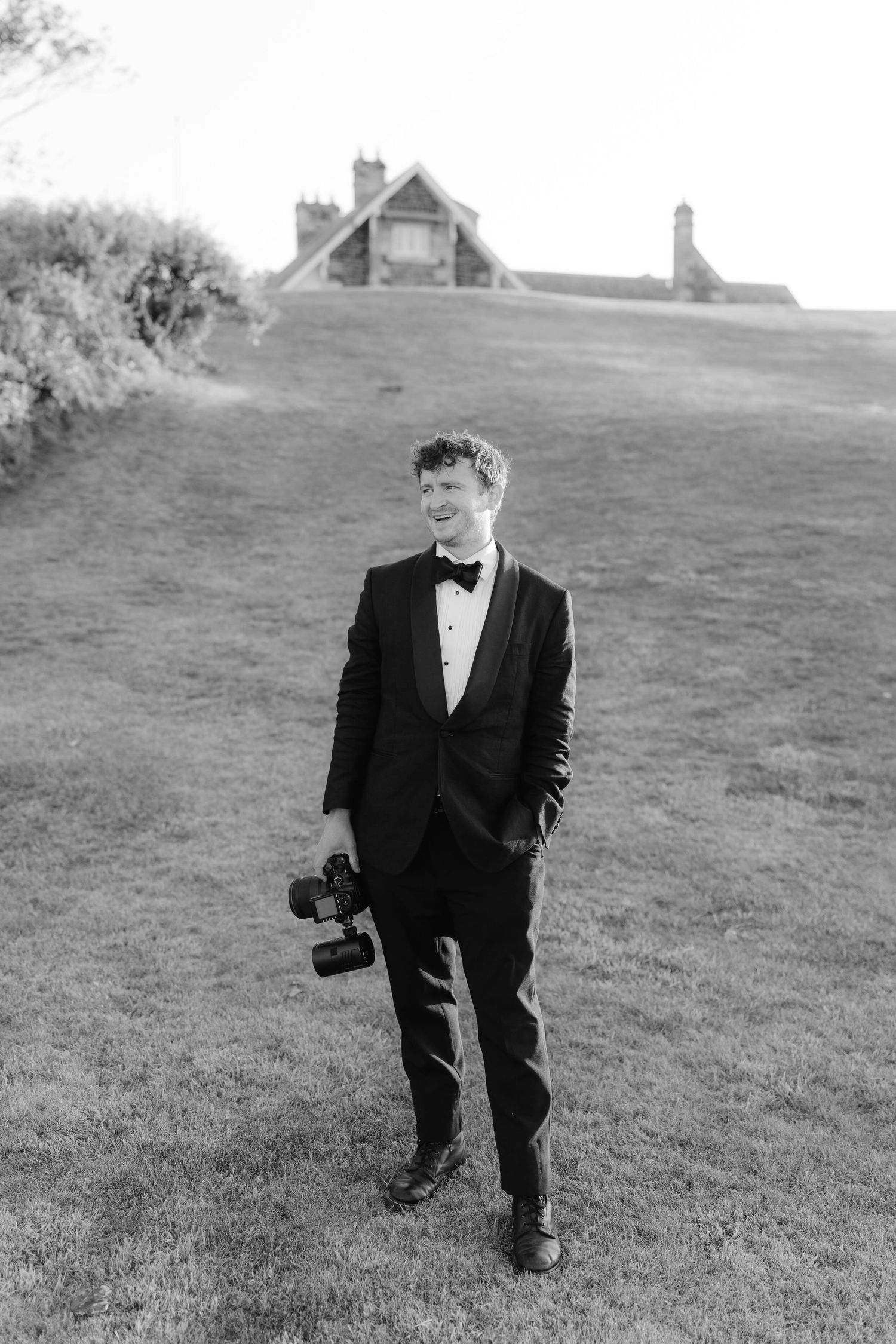 Black and white photo of a wedding photographer in a tuxedo holding a camera on a grassy hill.