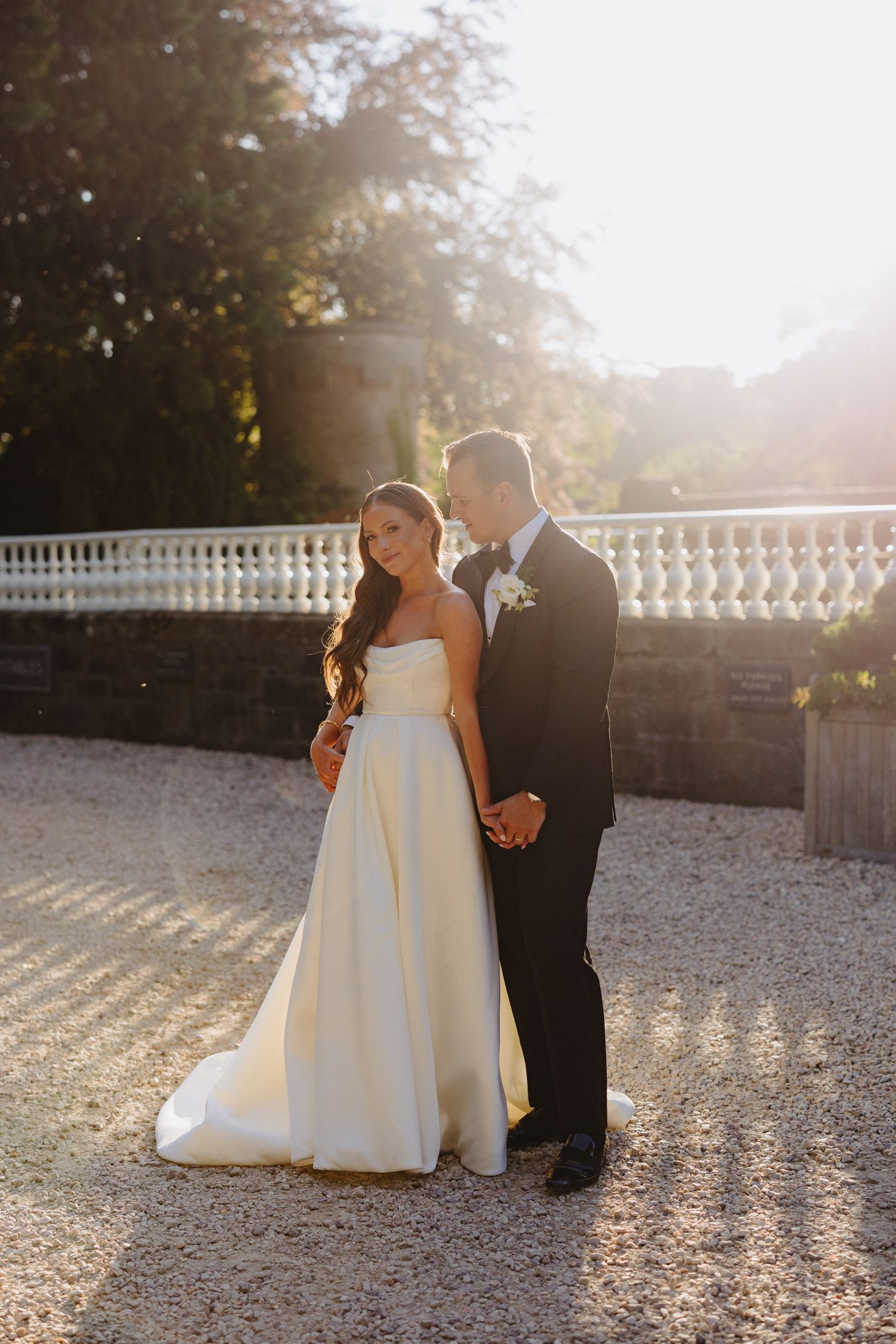Bride and groom holding hands in golden sunset light, bride in strapless ballgown at elegant gravel venue.