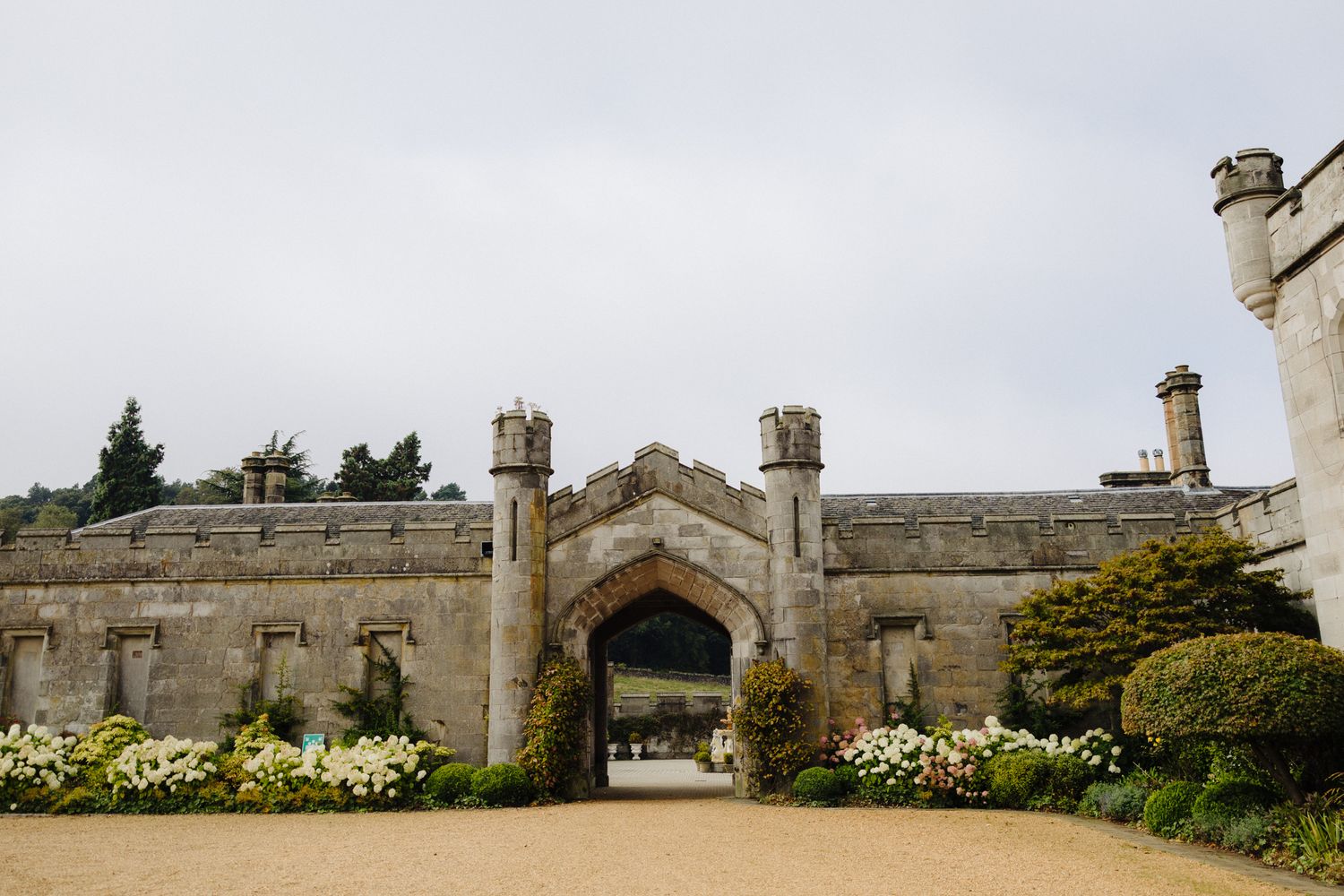 Historic stone gatehouse with Gothic arch entrance, flanked by turrets and white flowering shrubs under overcast skies.