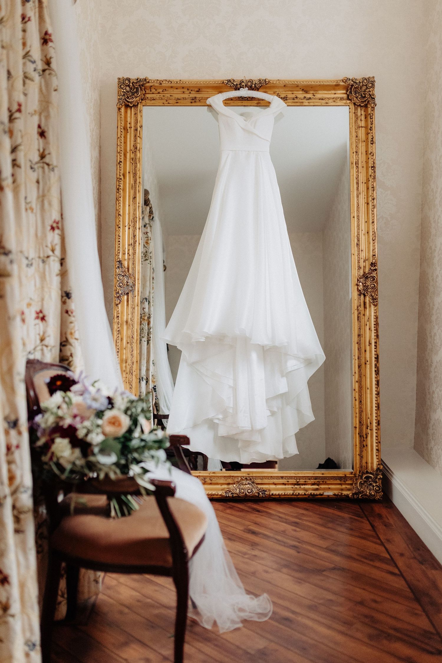 White wedding dress hanging on gold ornate mirror with bridal bouquet on vintage chair in elegant room.