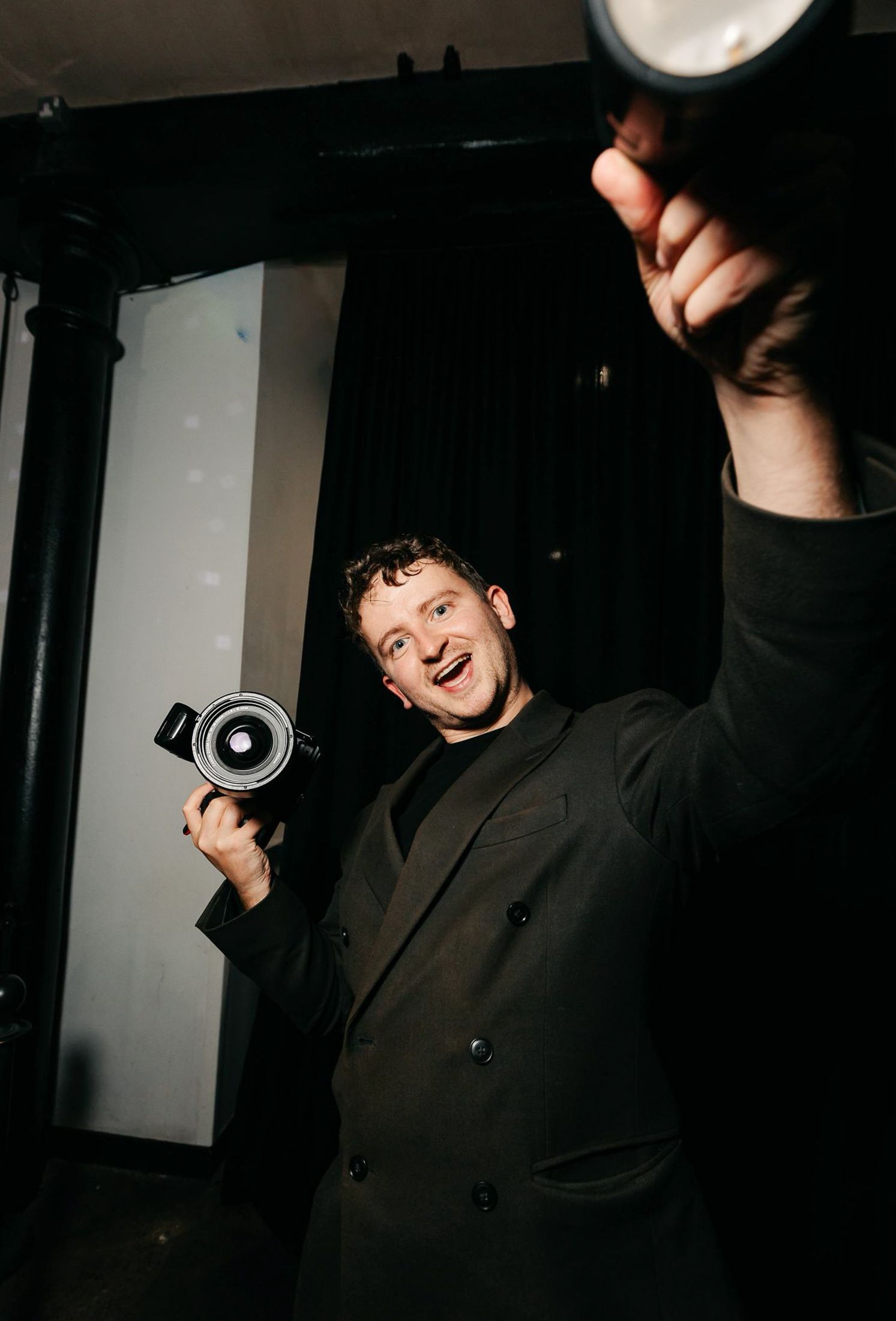 Man in black suit smiling while holding a vintage camera at an event, with studio lighting in background.