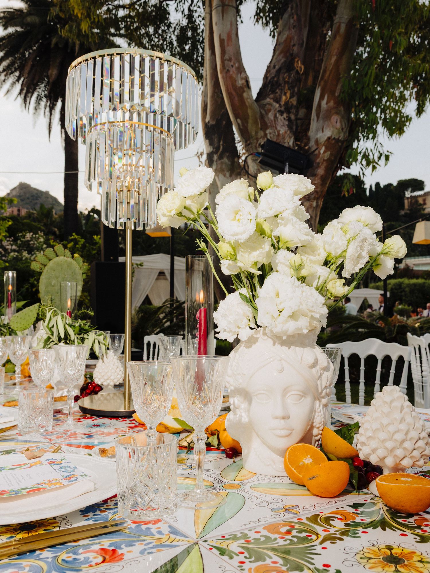 Elegant outdoor tablescape with crystal chandelier, white floral centerpiece in ceramic head vase, colorful linens, and fresh citrus fruits.