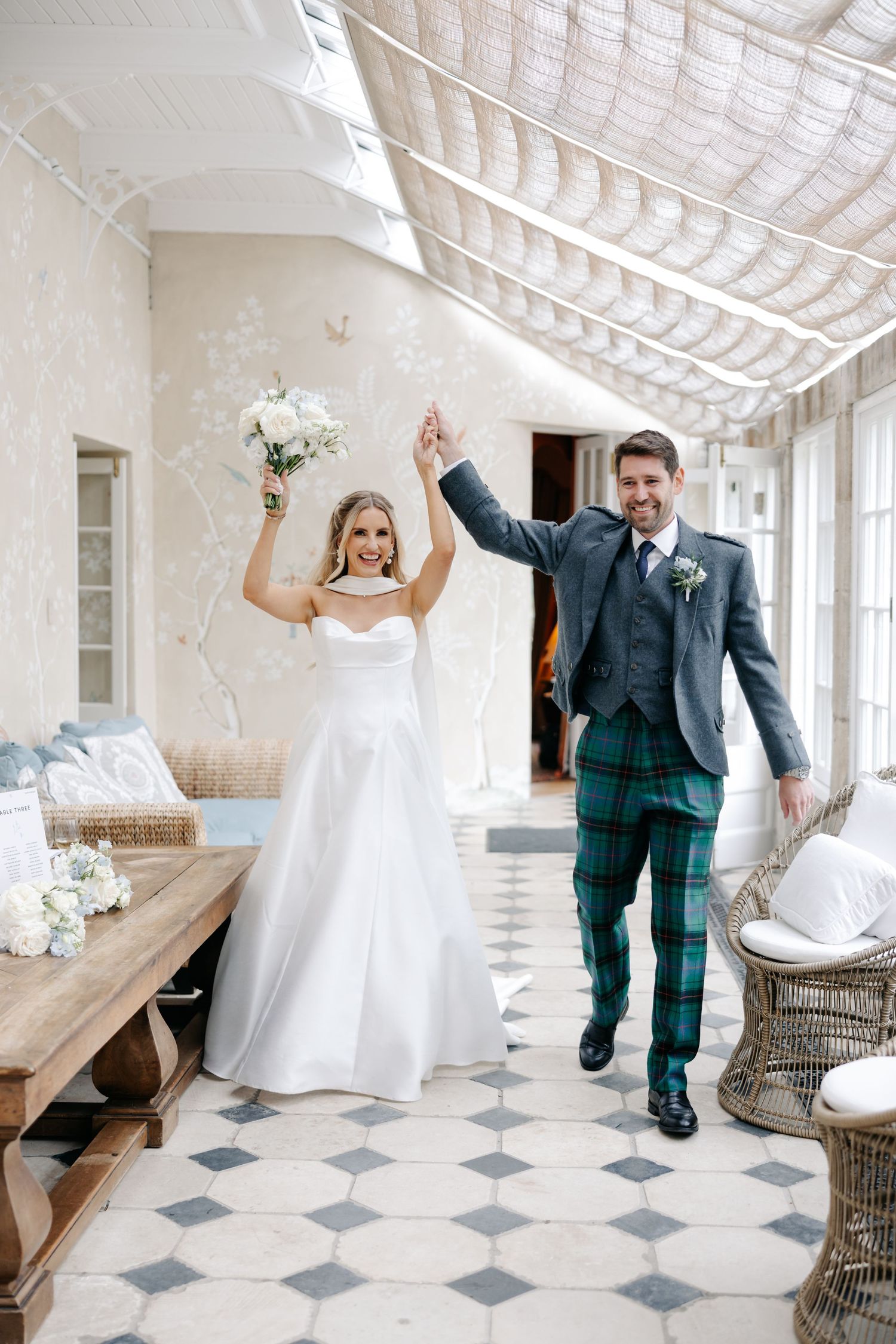 Joyful bride in white gown and groom in green tartan kilt raise hands in celebration walking through elegant venue.