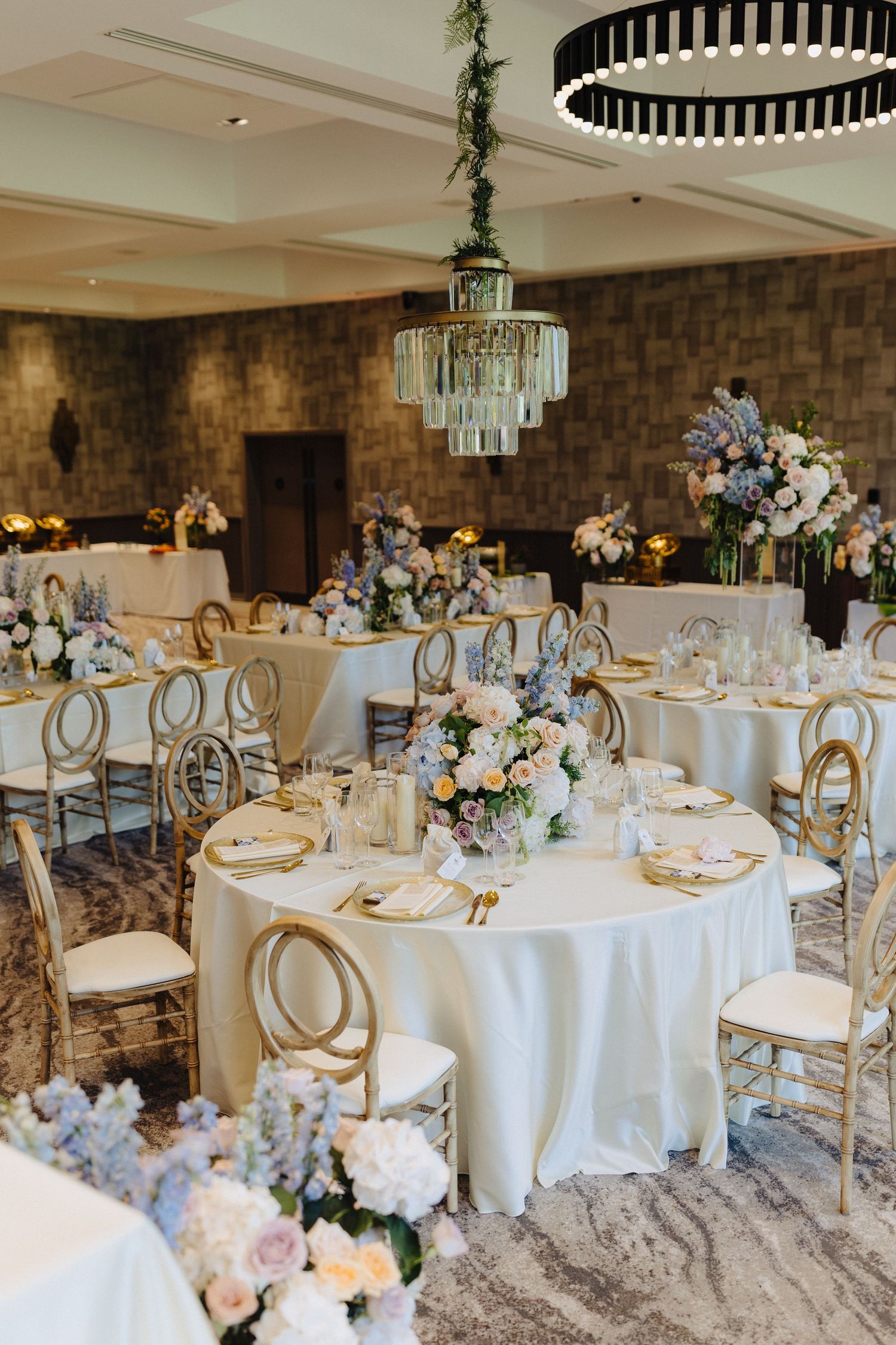 Elegant wedding reception hall with round tables, gold chiavari chairs, crystal chandeliers, and pastel floral centerpieces.