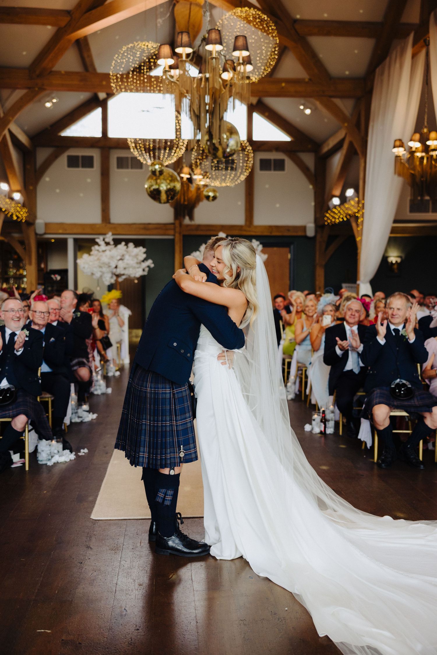 Newlyweds share first kiss under golden chandeliers as guests applaud in a rustic wedding venue.