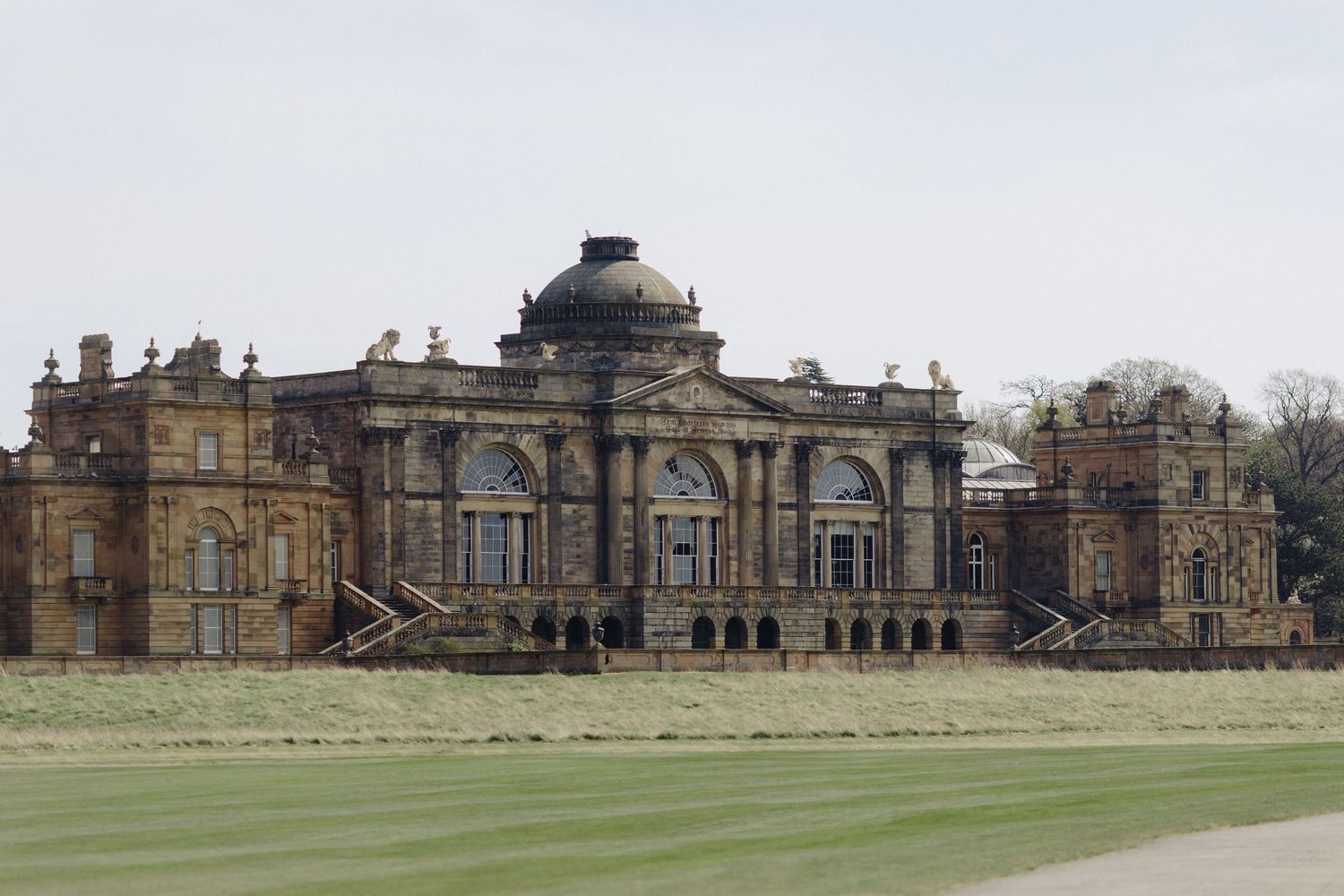 Grand historic English country mansion with ornate baroque architecture, dome, and manicured lawns under a pale sky.