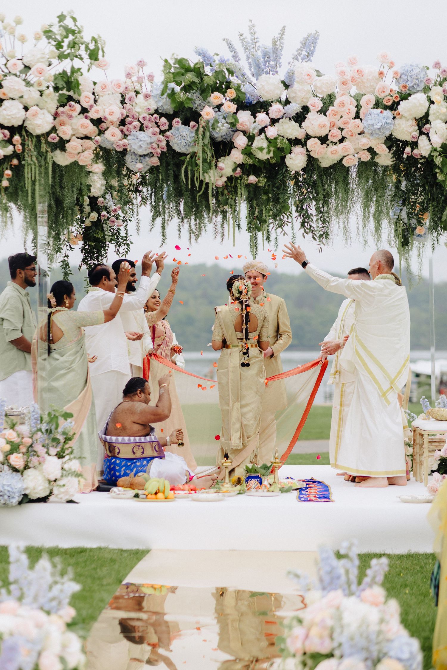 Outdoor Indian wedding ceremony under floral arch with pink blooms, guests tossing petals over couple in traditional attire.