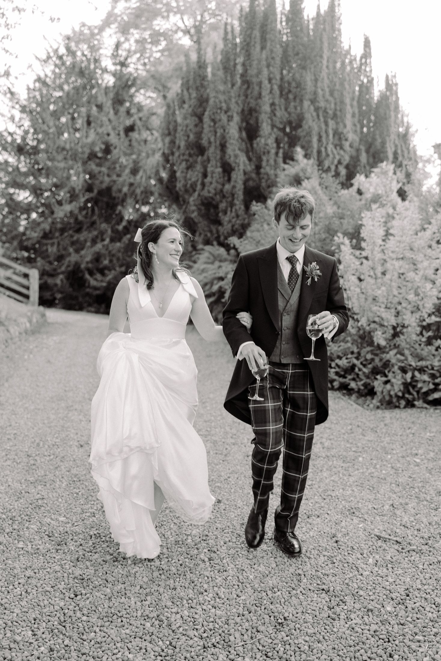 Black and white photo of a bride in white gown and groom in tartan kilt walking together holding champagne glasses.