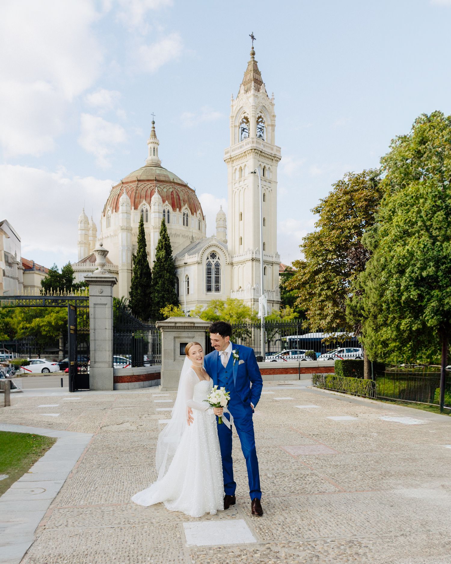 Bride and groom pose in front of a grand white church with a tall bell tower on their wedding day.