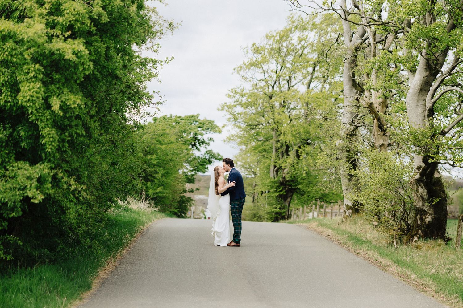 Bride and groom share a romantic kiss on a tree-lined country road on their wedding day.