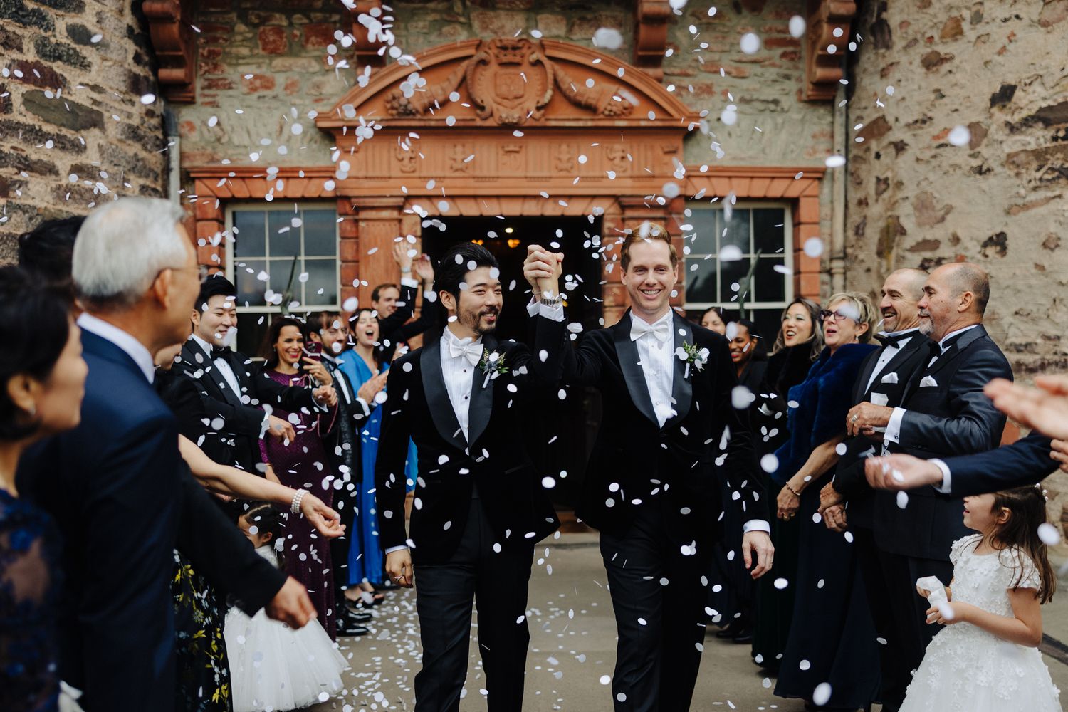 Two grooms celebrate their wedding exit through a confetti shower surrounded by cheering guests at a rustic stone venue.