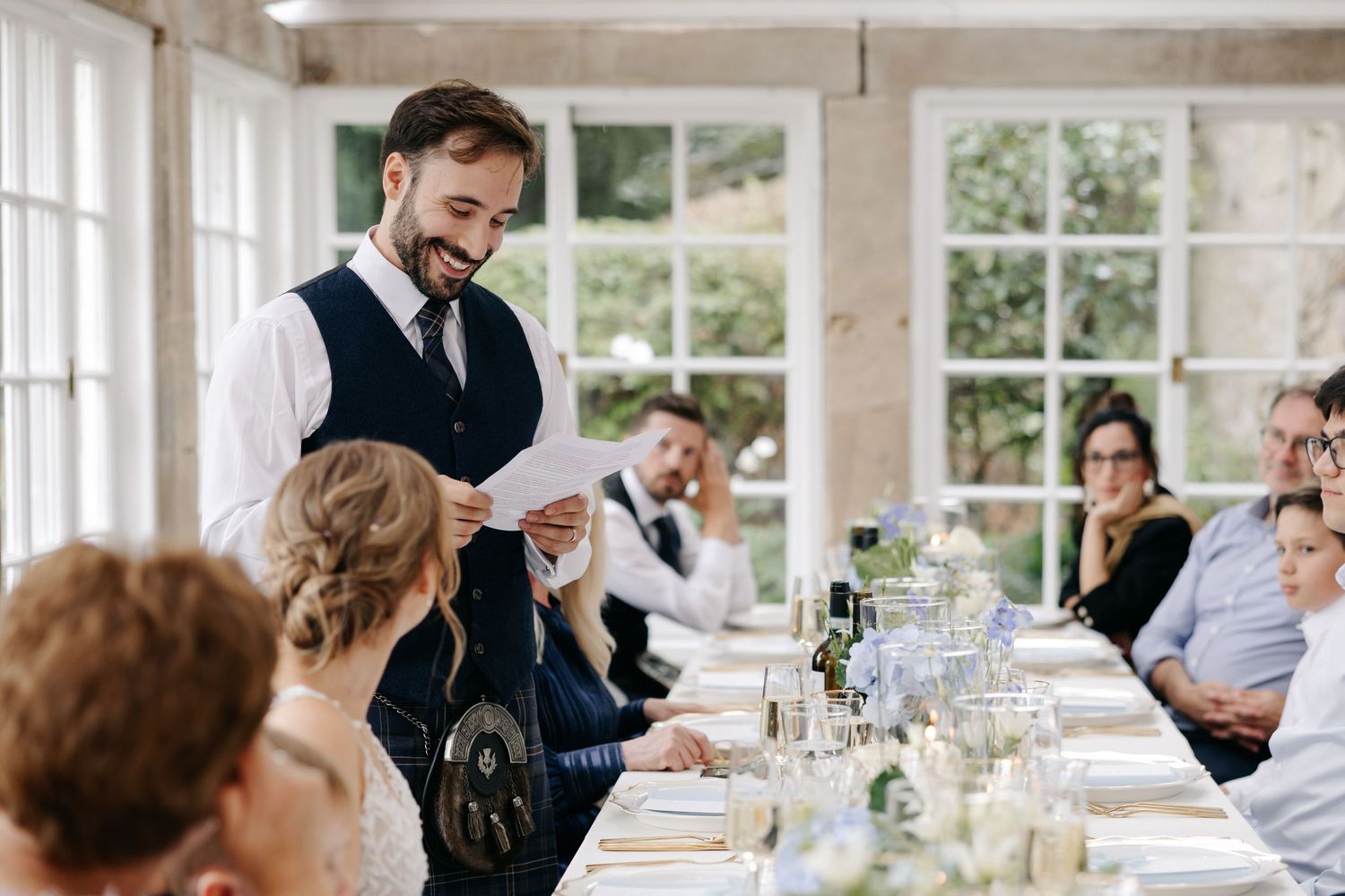 Man in vest reads speech at elegant wedding reception dinner table with guests listening attentively.