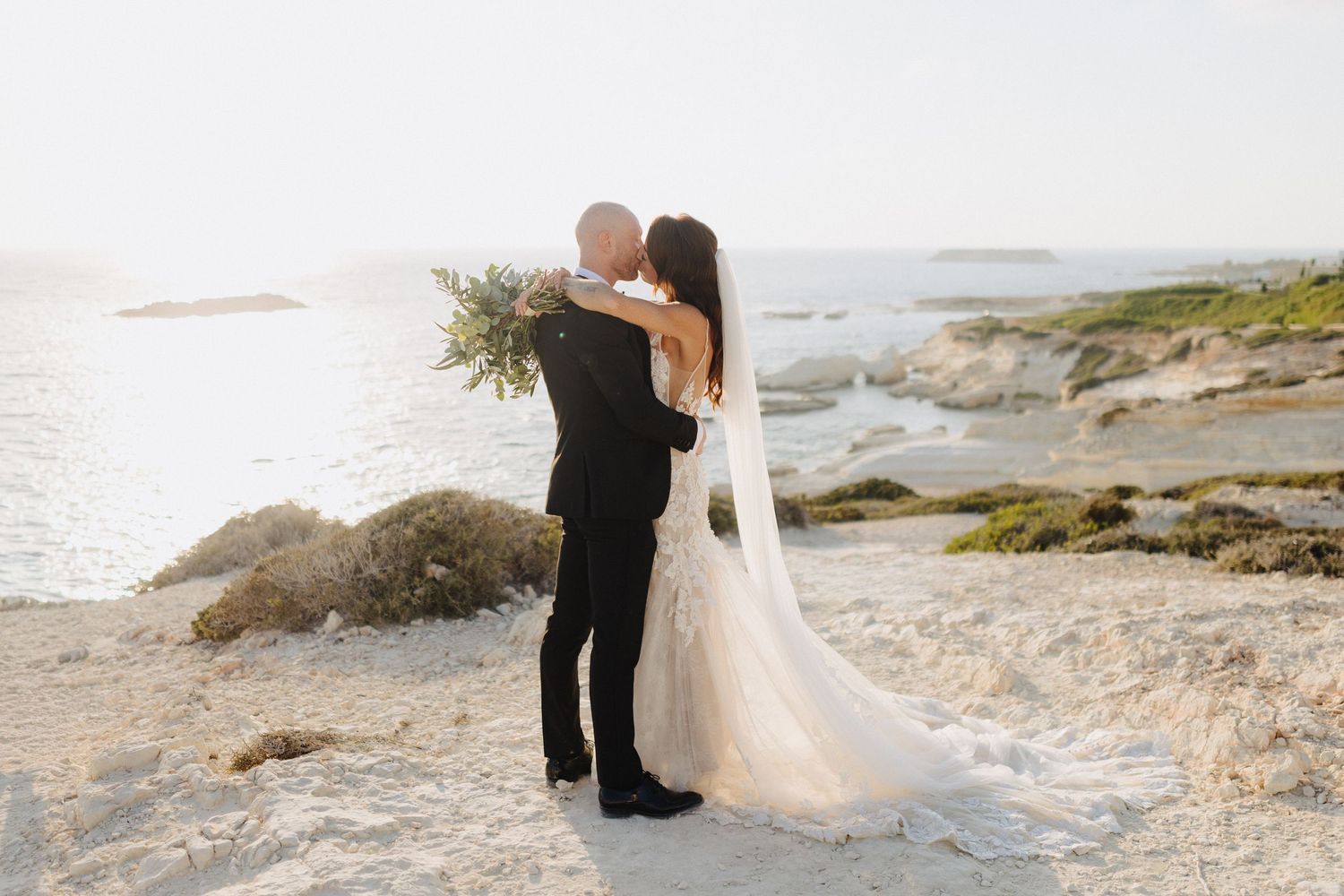 Romantic couple sharing a kiss on a rocky coastal landscape at golden hour with ocean views.