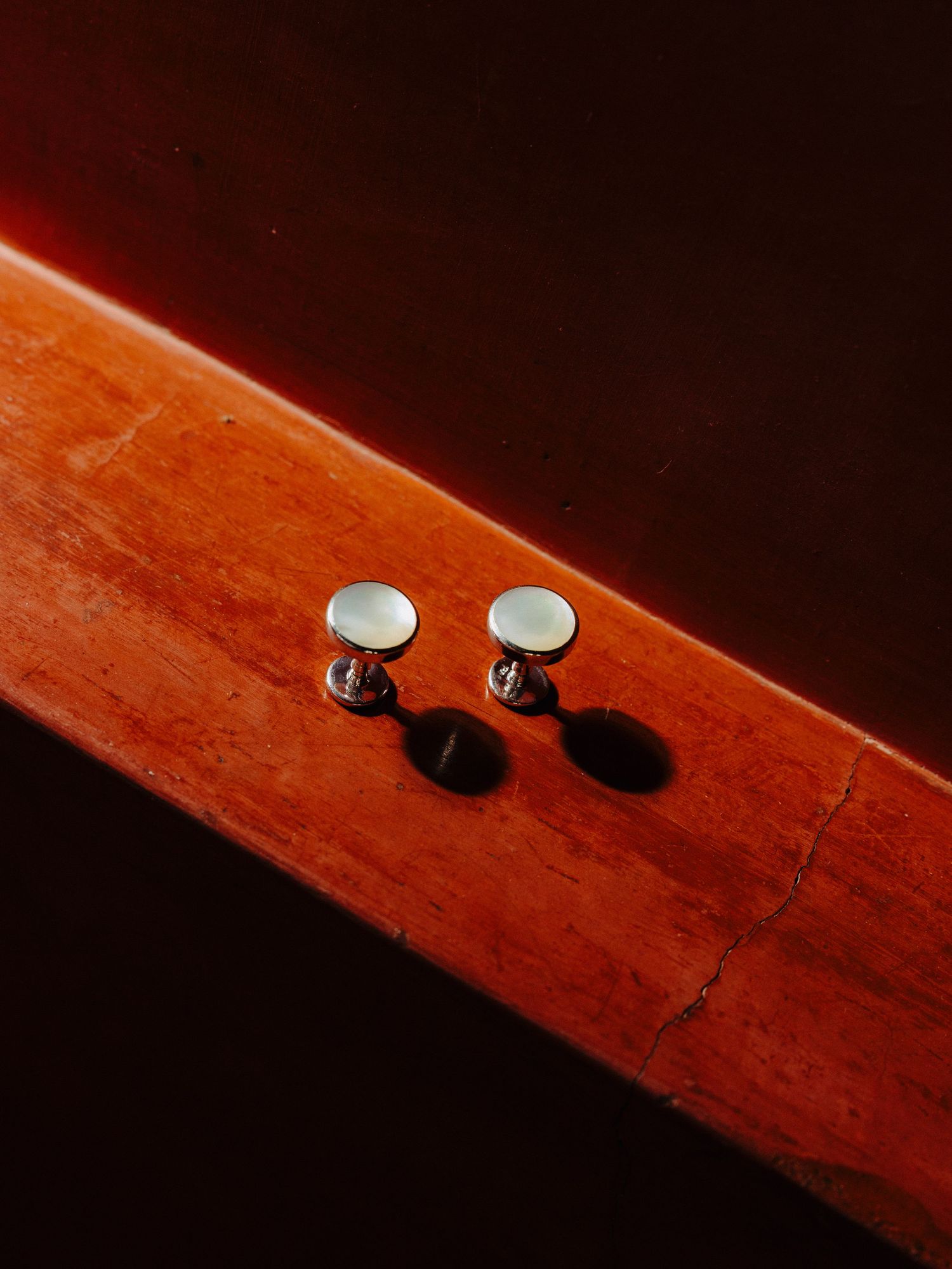 Two silver tuxedo shirt studs resting on warm reddish-brown wooden surface with dramatic lighting.