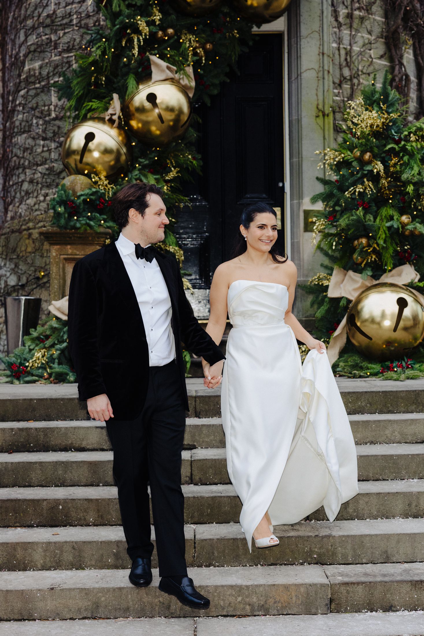 Bride and groom holding hands walking down stone steps with festive gold bell Christmas decorations.