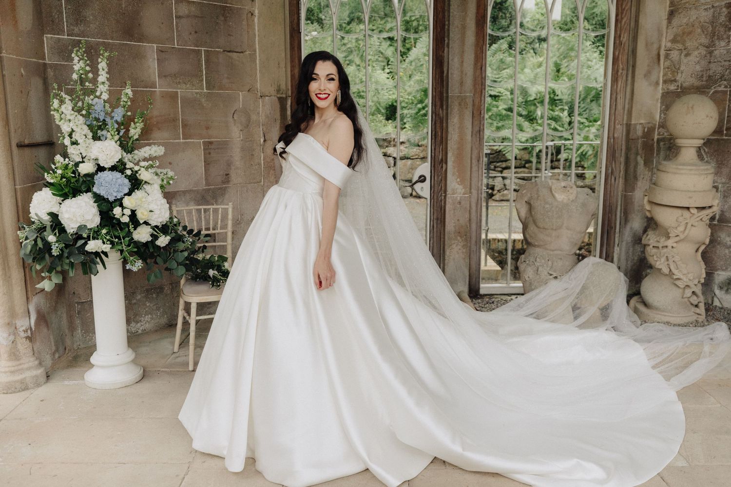 Bride in elegant off-shoulder white ball gown with long train poses in a stone venue with floral arrangements.