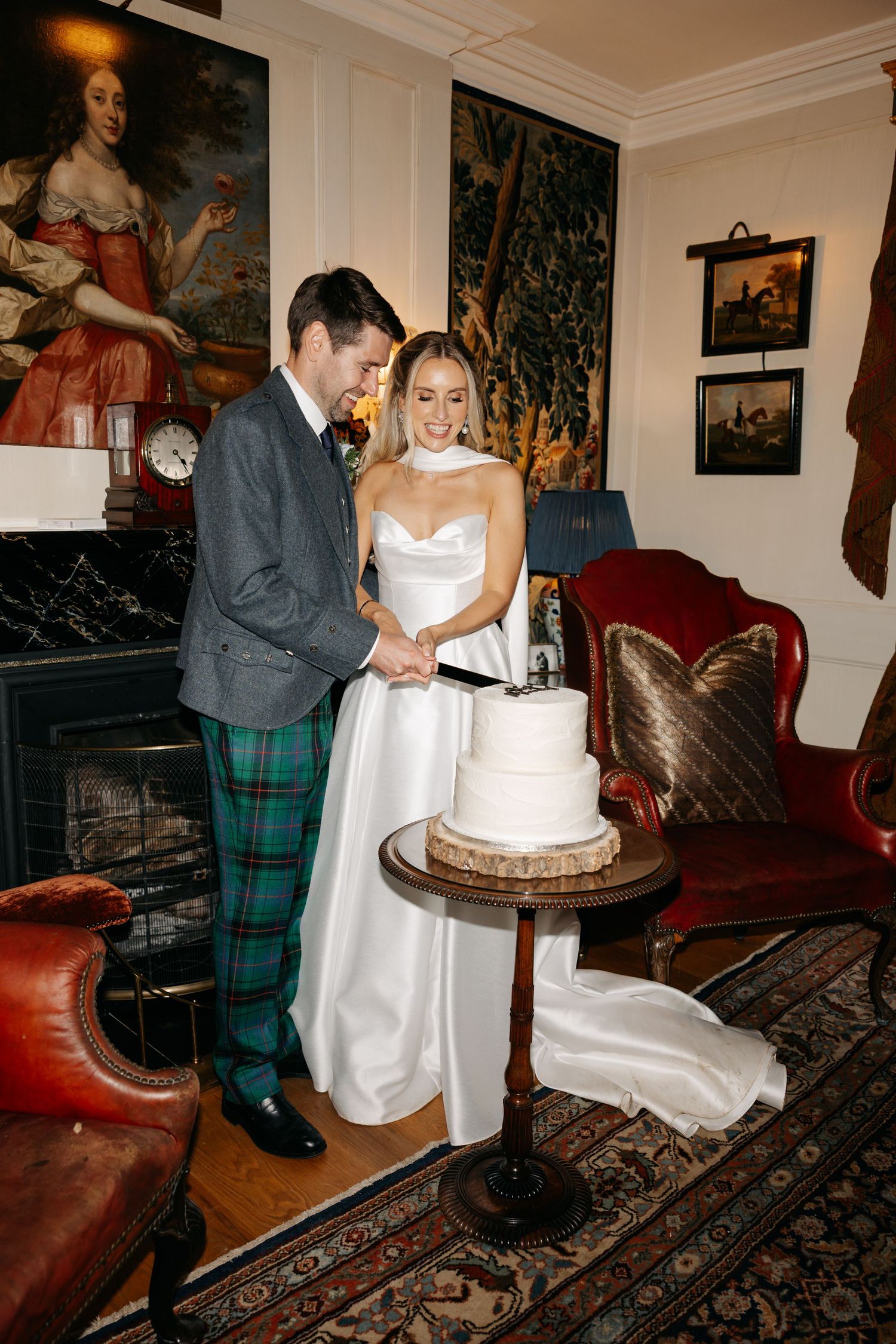 Bride and groom cutting a white tiered wedding cake in an elegant room with tartan trousers and a satin wedding gown.
