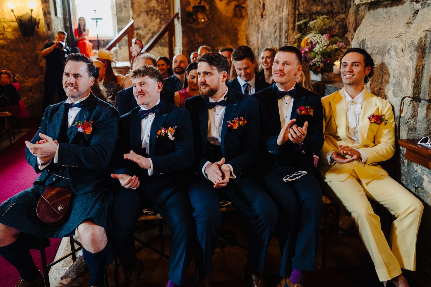 Groomsmen in navy suits with floral buttonholes laughing during a wedding ceremony in a rustic stone venue.