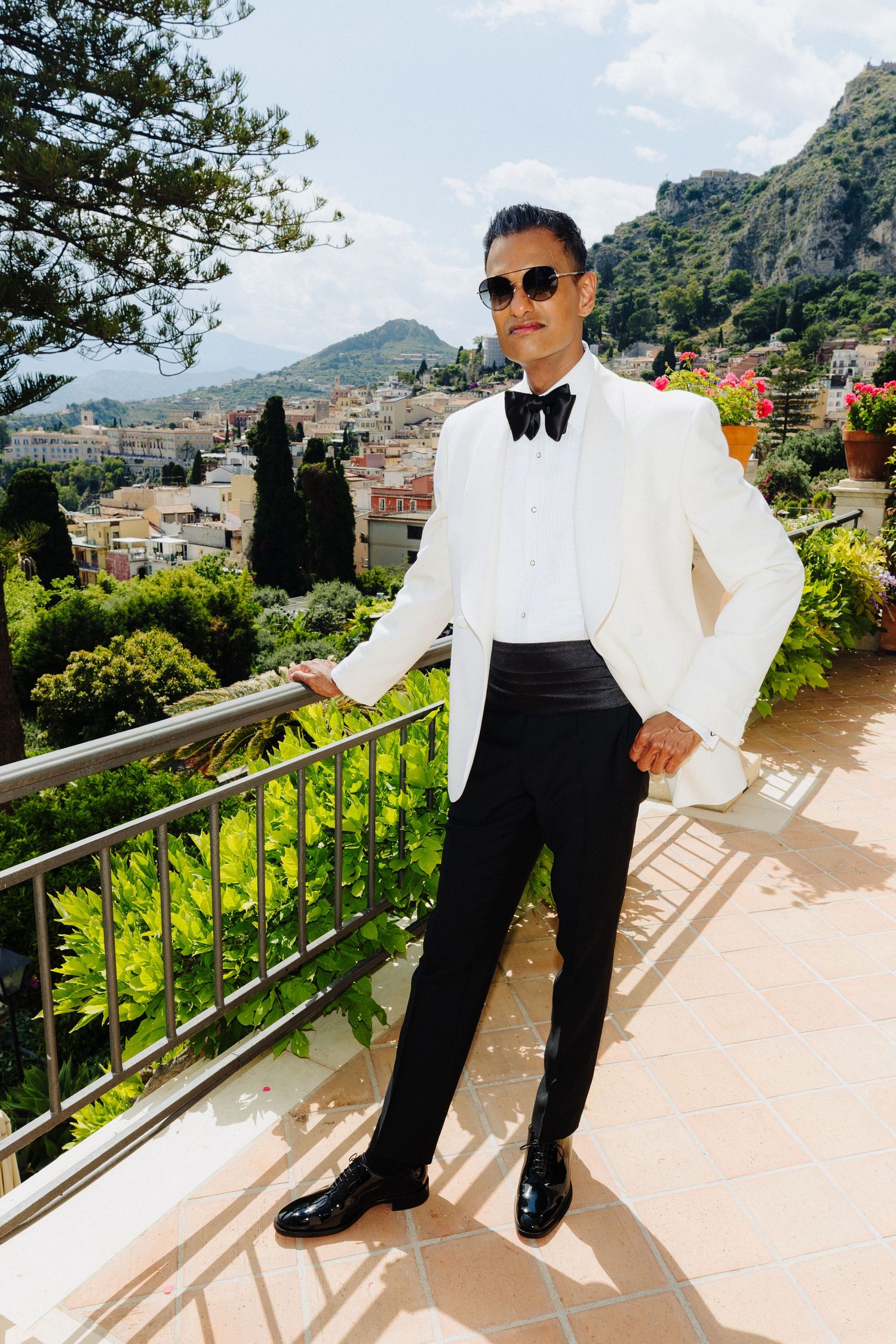 Groom in white tuxedo jacket and black bow tie posing on scenic Italian terrace overlooking Taormina.