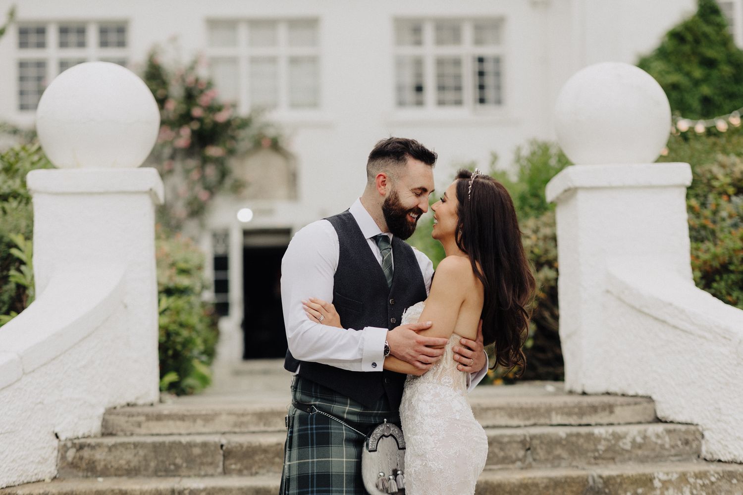Bride and groom sharing romantic moment on white manor steps, groom in Scottish tartan kilt.
