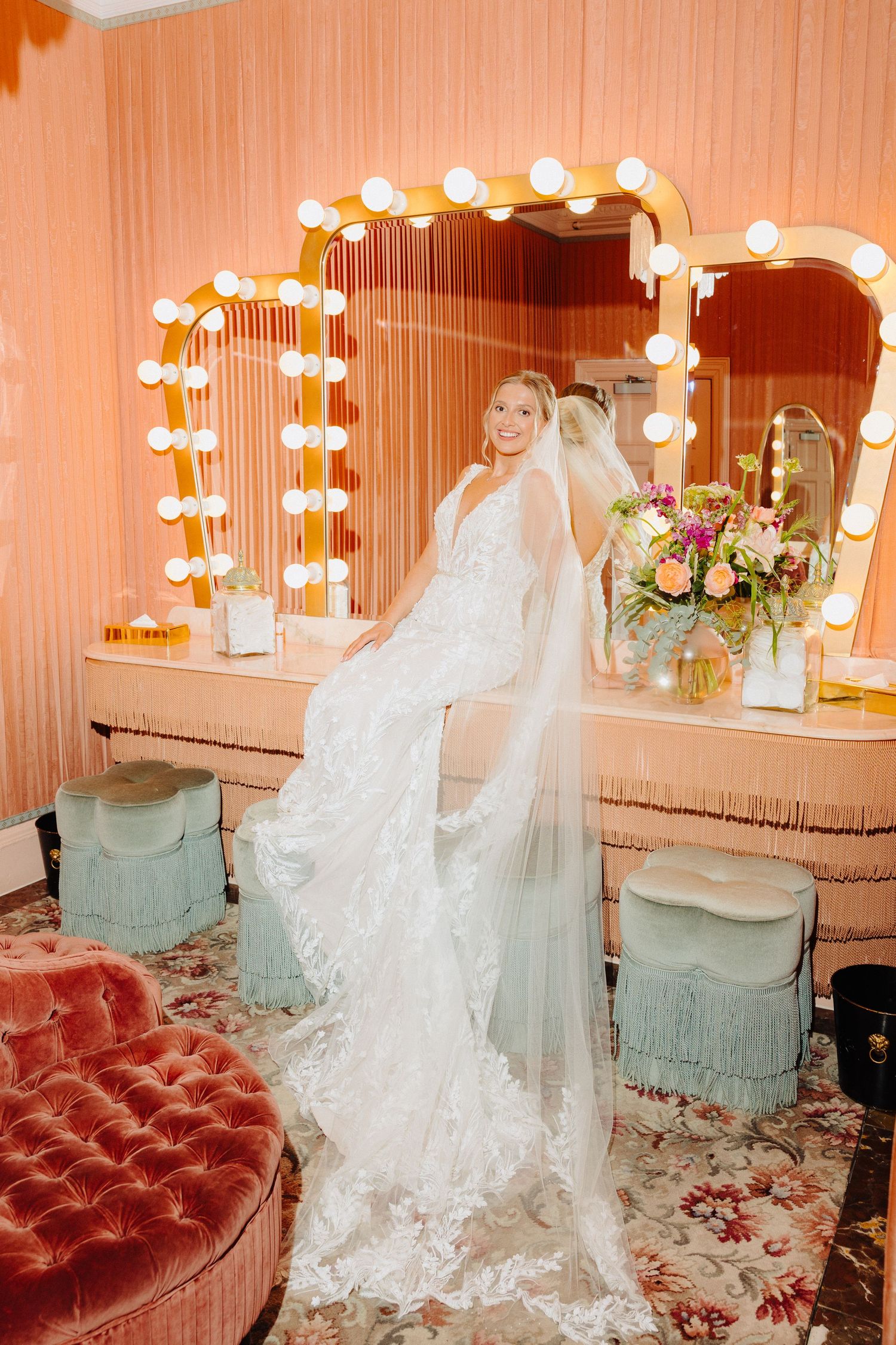 Radiant bride in a lace gown and veil posing before a glamorous Hollywood vanity mirror.