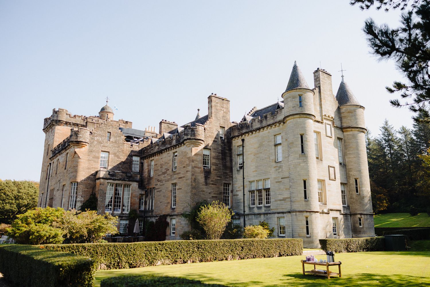 A grand Scottish castle with turrets and stone facade surrounded by manicured lawns and trees under a clear blue sky.