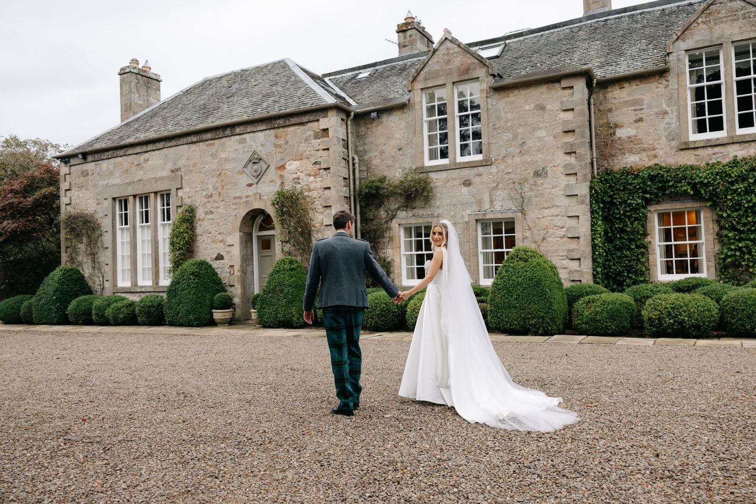 Bride and groom holding hands walking toward a grand stone manor house surrounded by manicured hedges on their wedding day.