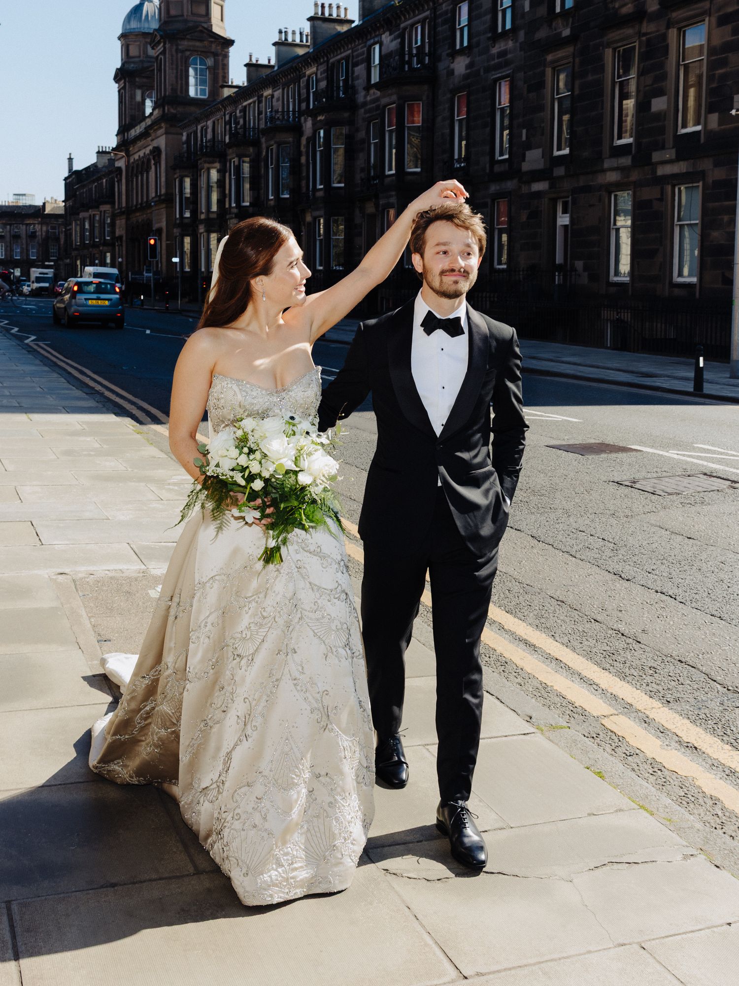 Stylish bride twirling groom on a sunny Edinburgh street, wearing a lace gown and tuxedo.