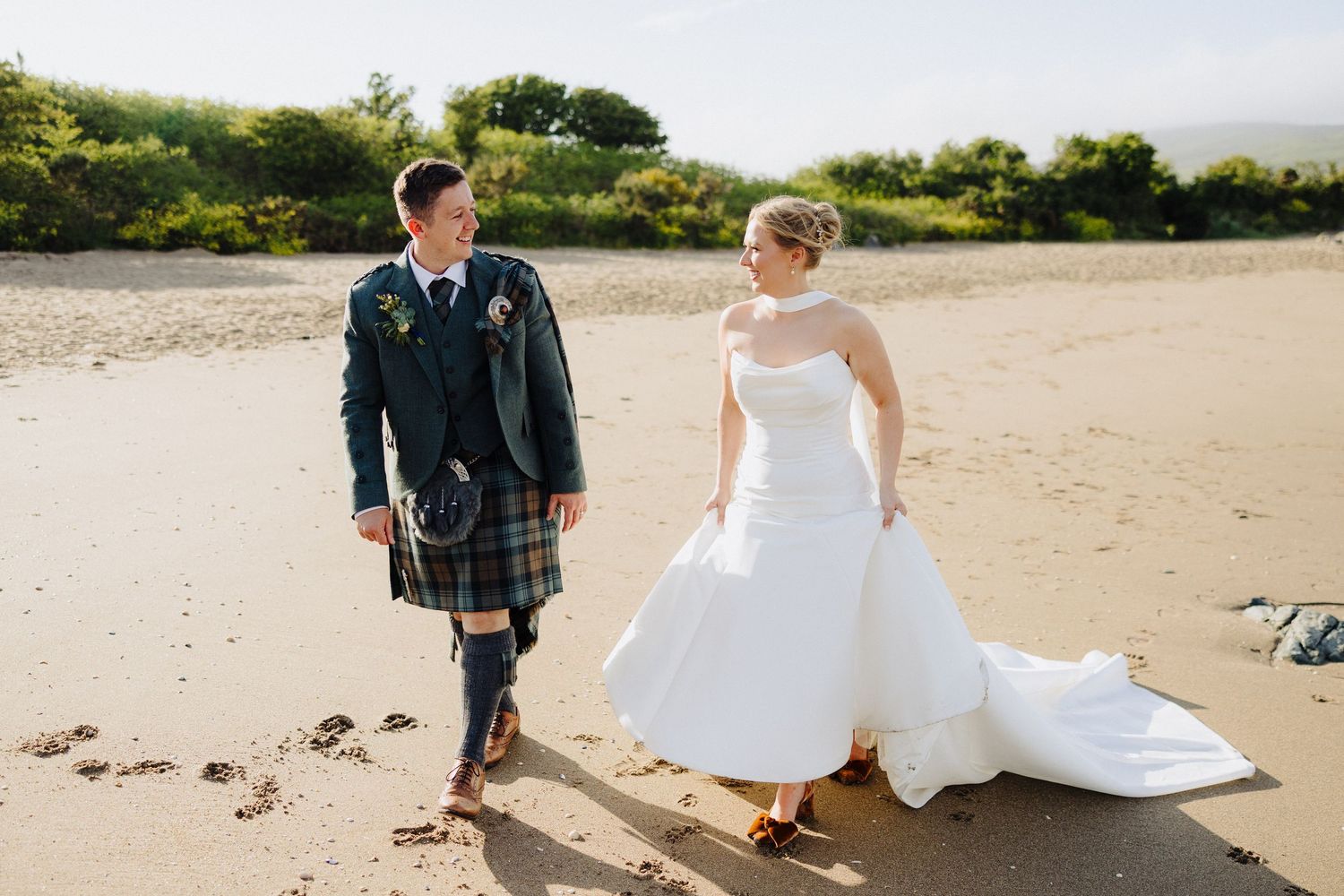 Bride and groom walking on sandy beach, groom wearing Scottish tartan kilt, bride in elegant white wedding gown.
