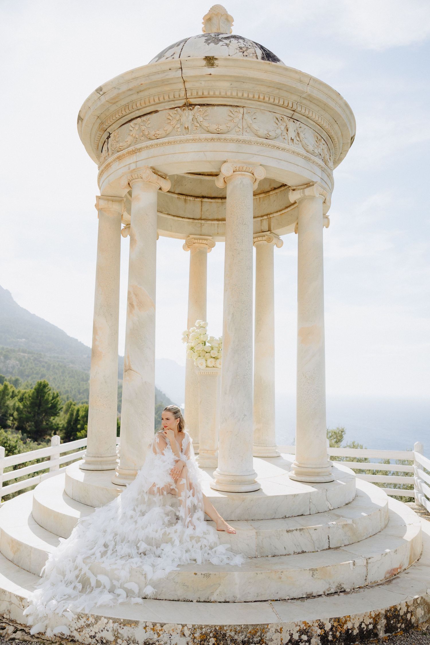 Bride in a flowing white feathered gown sits at a classical marble rotunda with columns and mountain views.