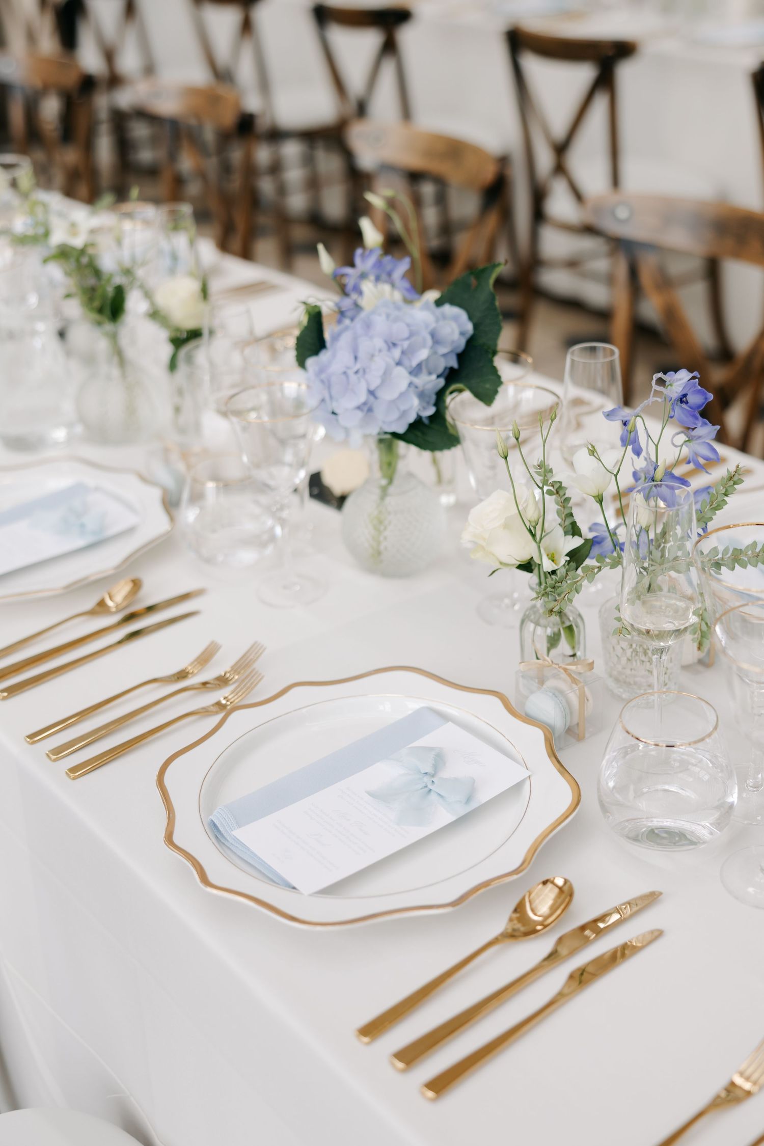 Elegant wedding table setting with gold cutlery, white plates, and blue hydrangea floral arrangements on white linen.