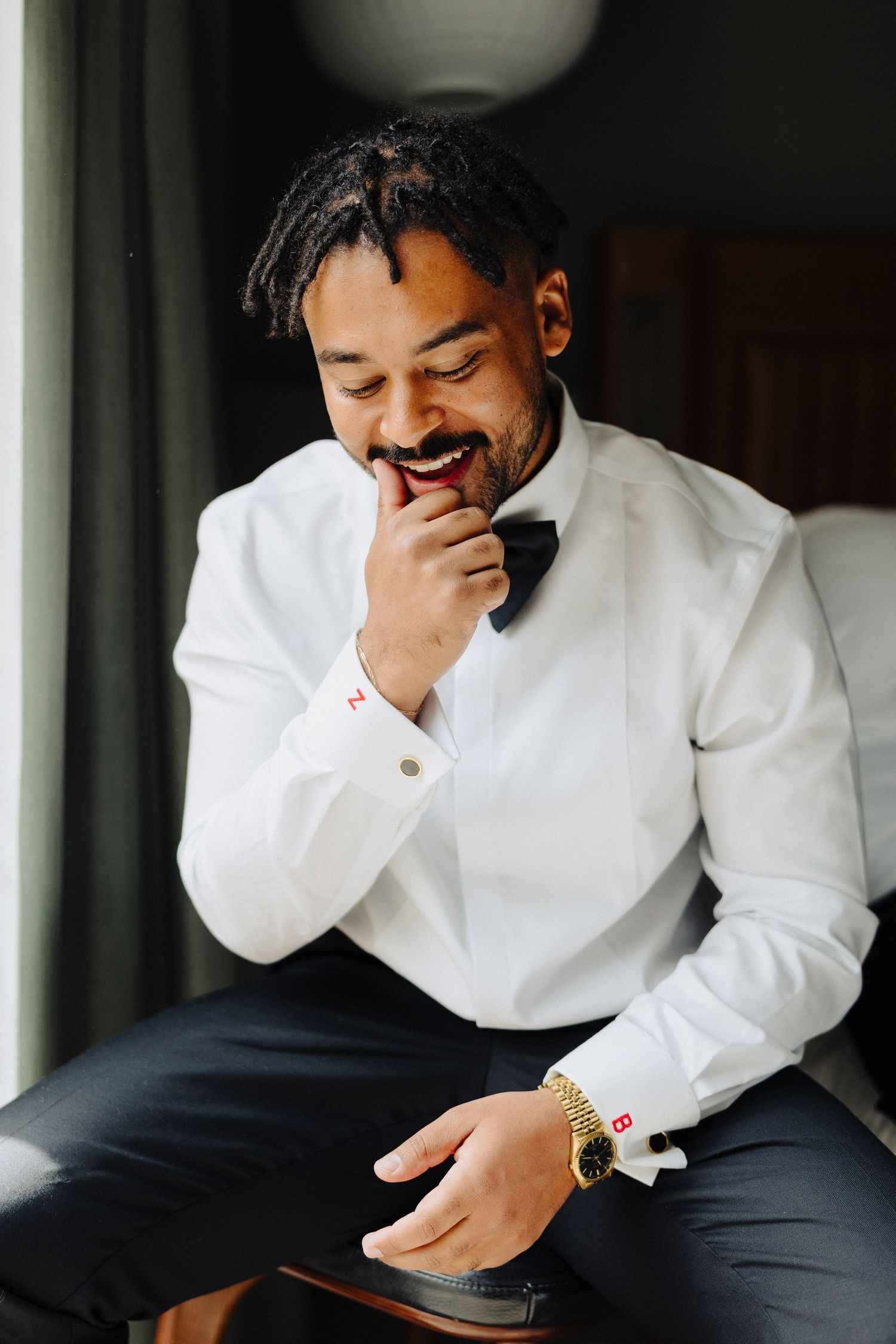 Smiling groom in white dress shirt and bow tie adjusts cufflinks while seated by a window.