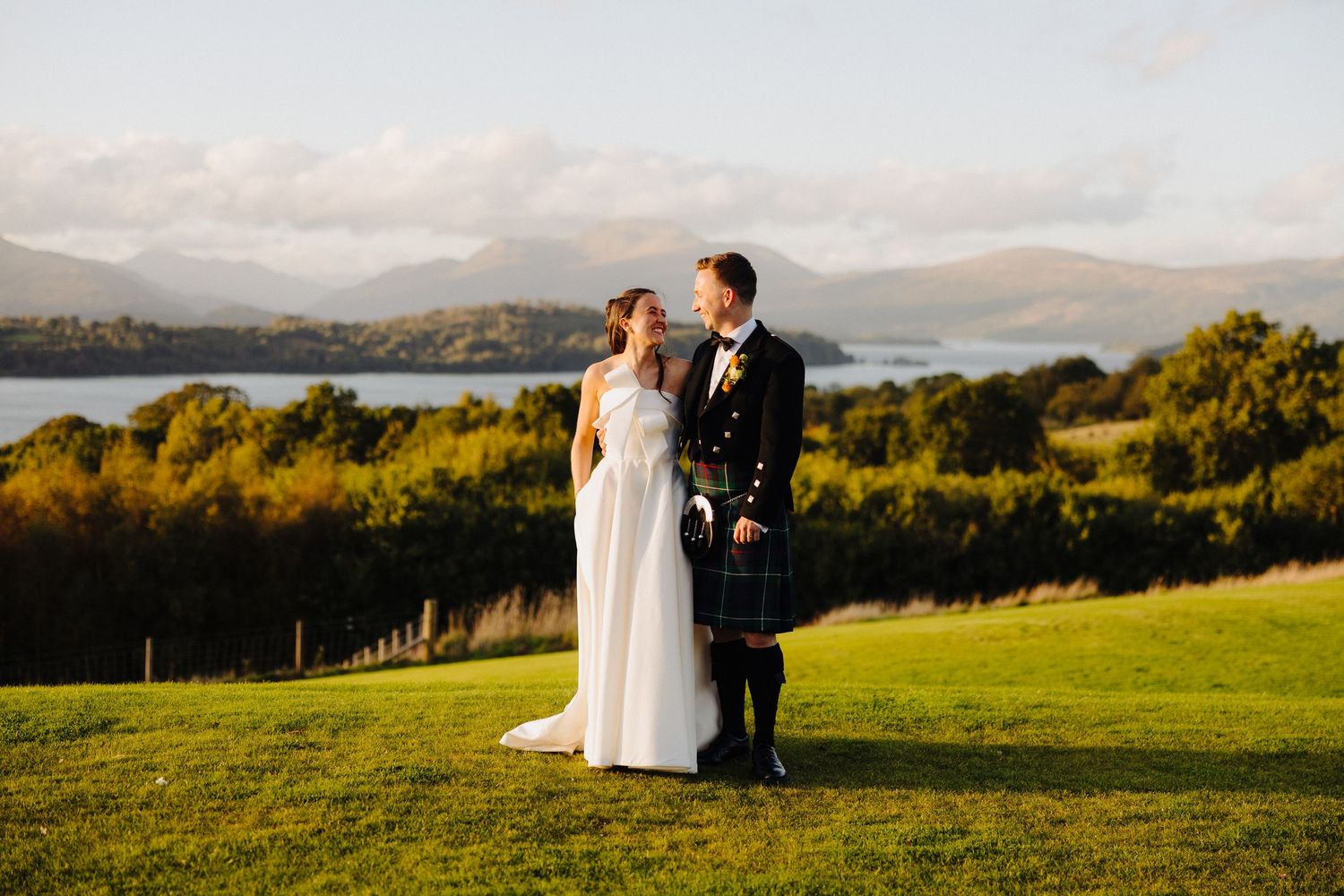 A bride and groom stand together on a green hillside in Scotland, with a loch and mountains in the golden evening light.