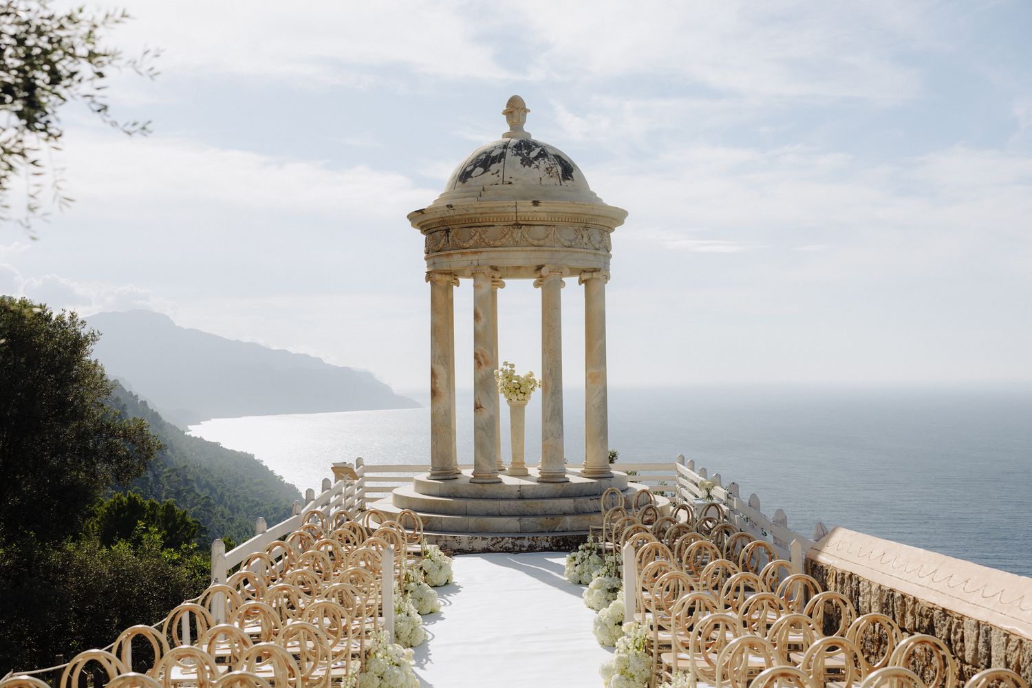 Elegant outdoor wedding ceremony at a cliffside temple with ocean views, white aisle, and gold chairs in Mallorca.