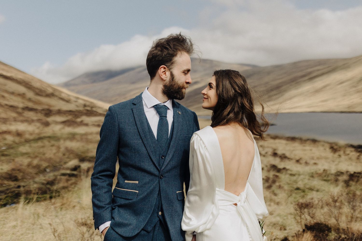 A couple in wedding attire shares a romantic moment in a scenic Scottish highland landscape with mountains and a loch.