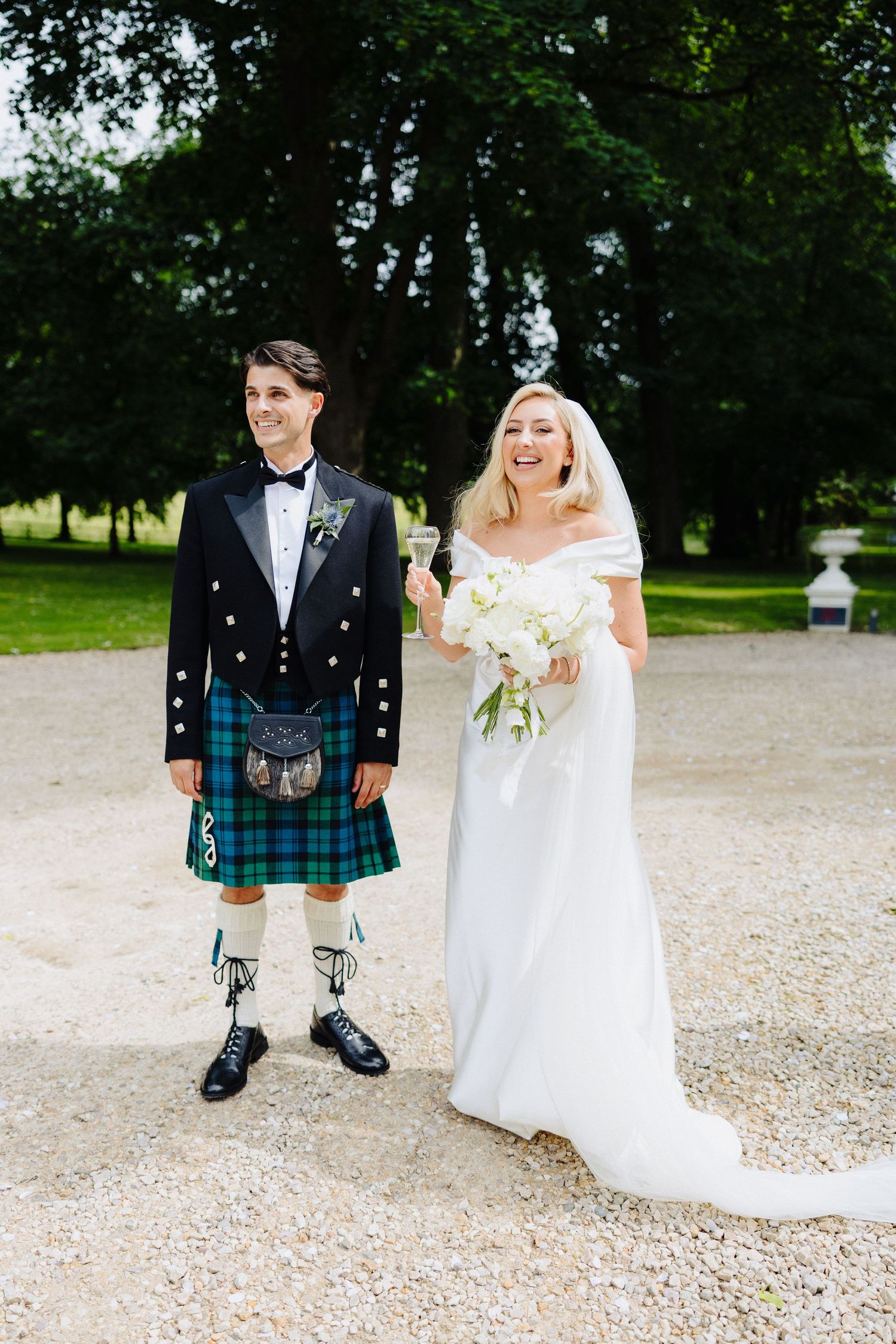 Bride in elegant white gown and groom in traditional Scottish kilt stand together on a gravel path.