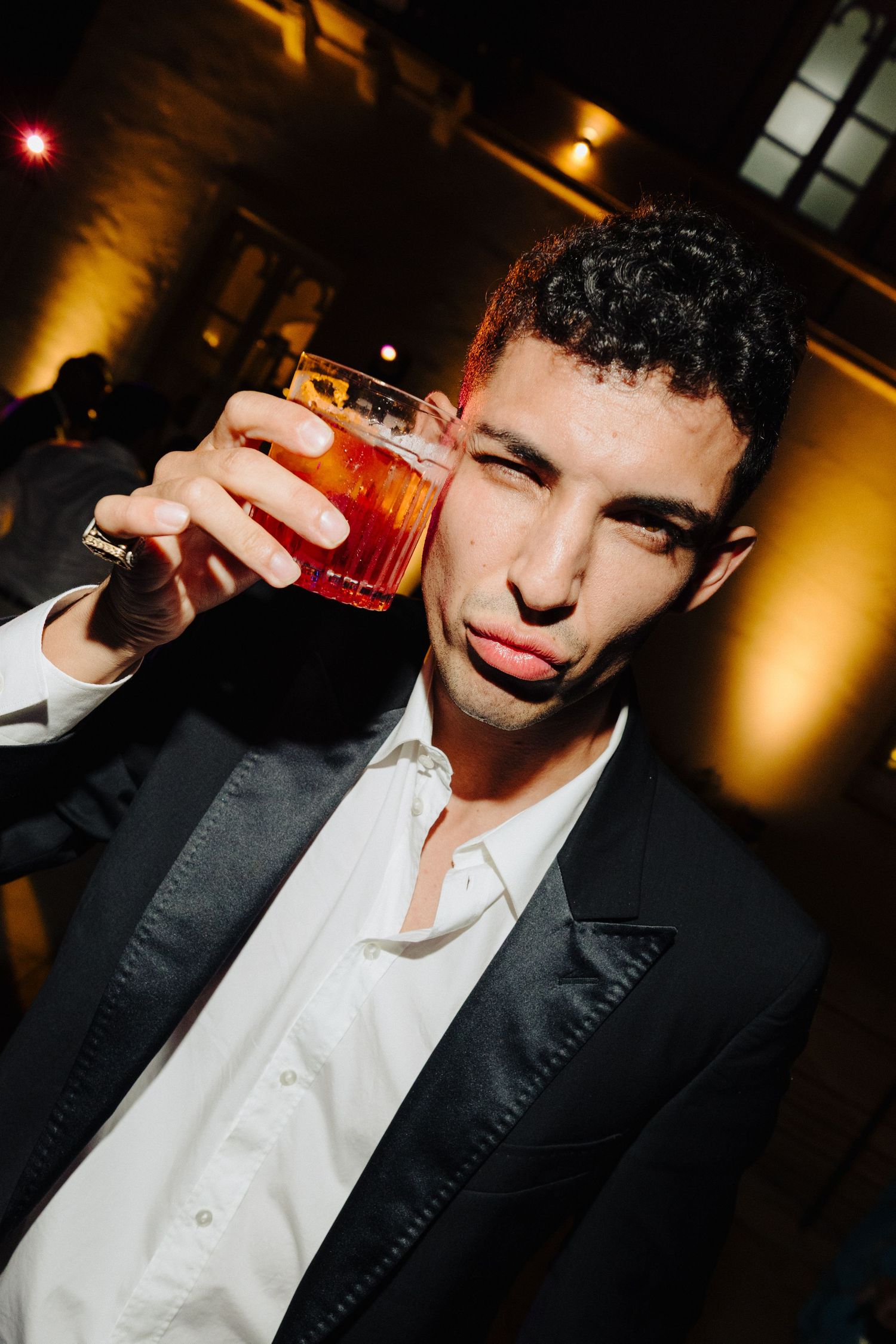 Stylish wedding guest in a black suit holding an amber cocktail drink at an evening indoor reception.
