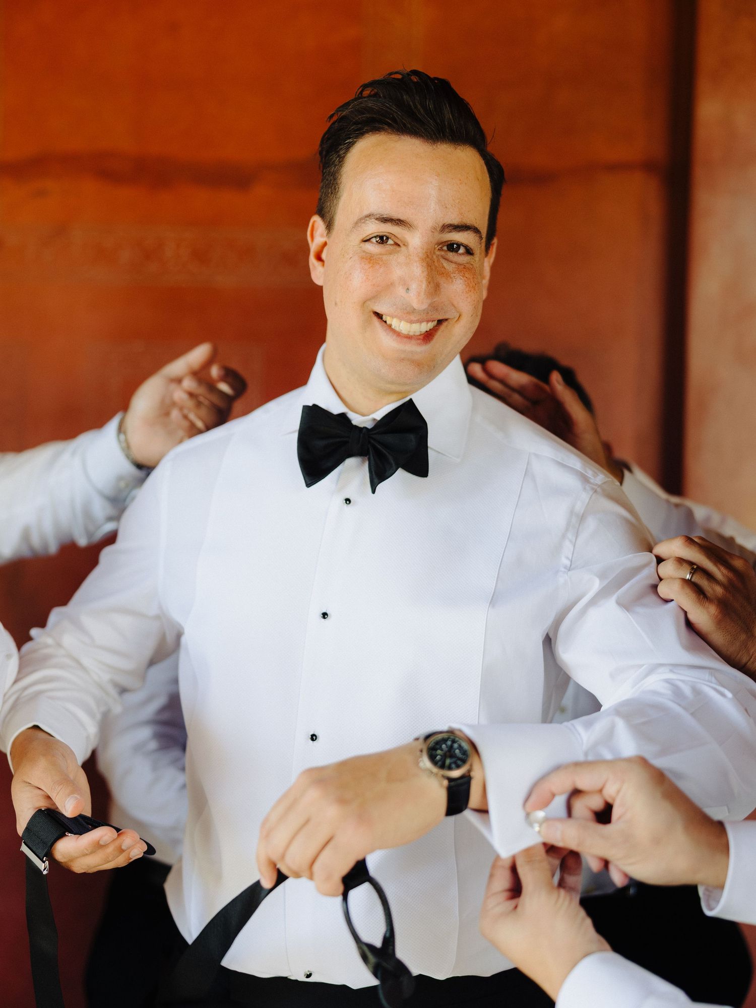 Smiling groom in white tuxedo shirt and black bow tie being helped by groomsmen while getting ready.