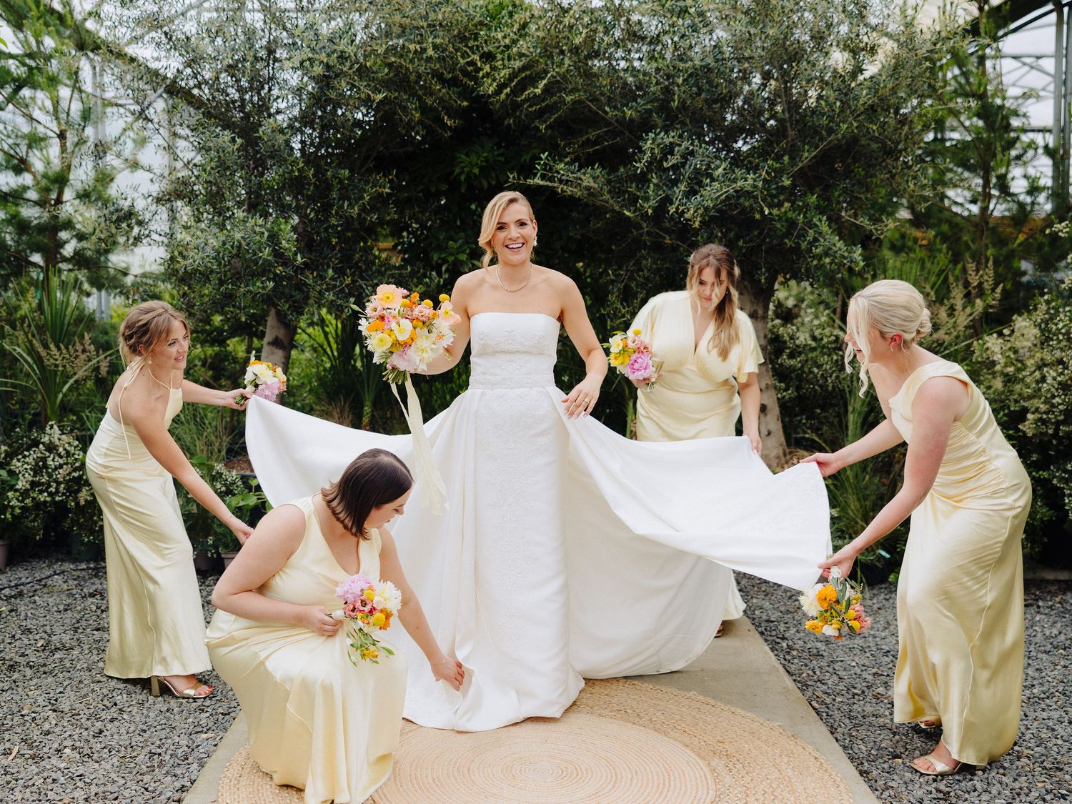 Bride in white gown surrounded by four bridesmaids in yellow dresses holding colorful bouquets in a garden setting.