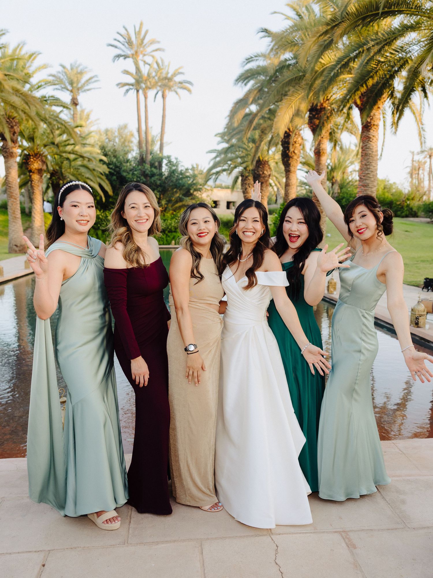 Bride with bridesmaids in mismatched colorful gowns posing joyfully in front of tall palm trees.